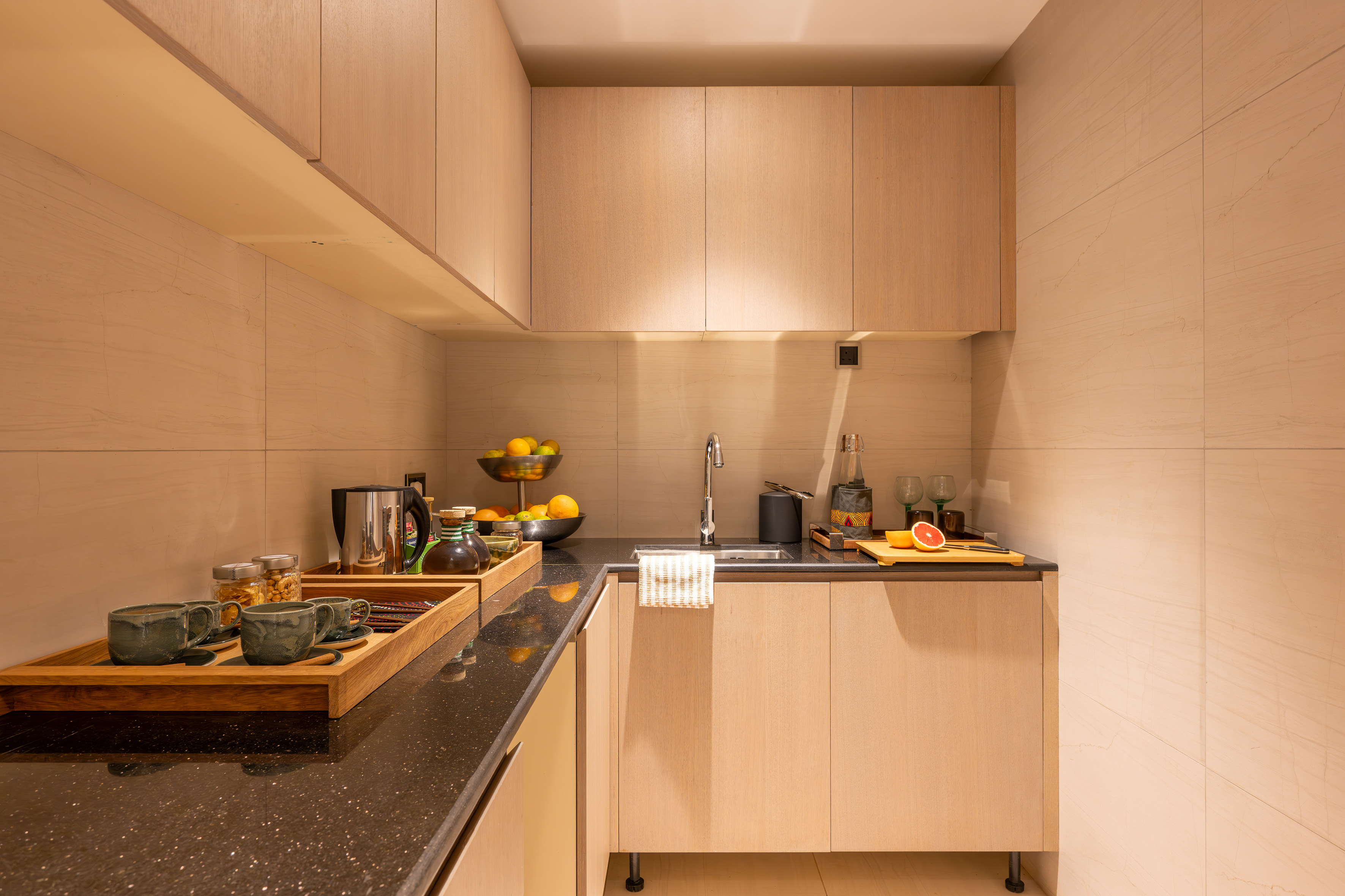 Modern kitchenette with light wood cabinets, dark counter, sink, coffee, and fruit.