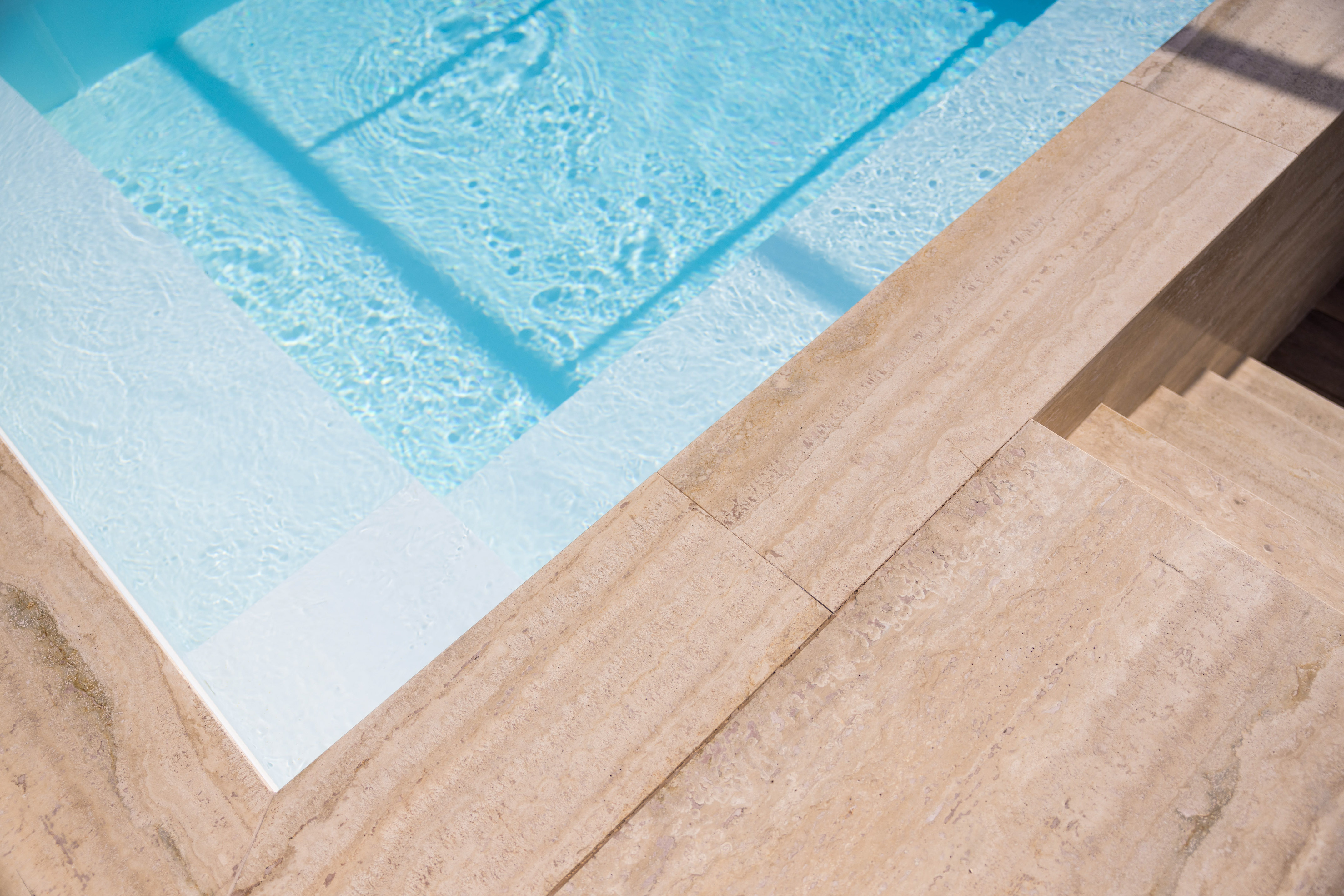 Clear blue swimming pool with white interior steps and beige stone coping.