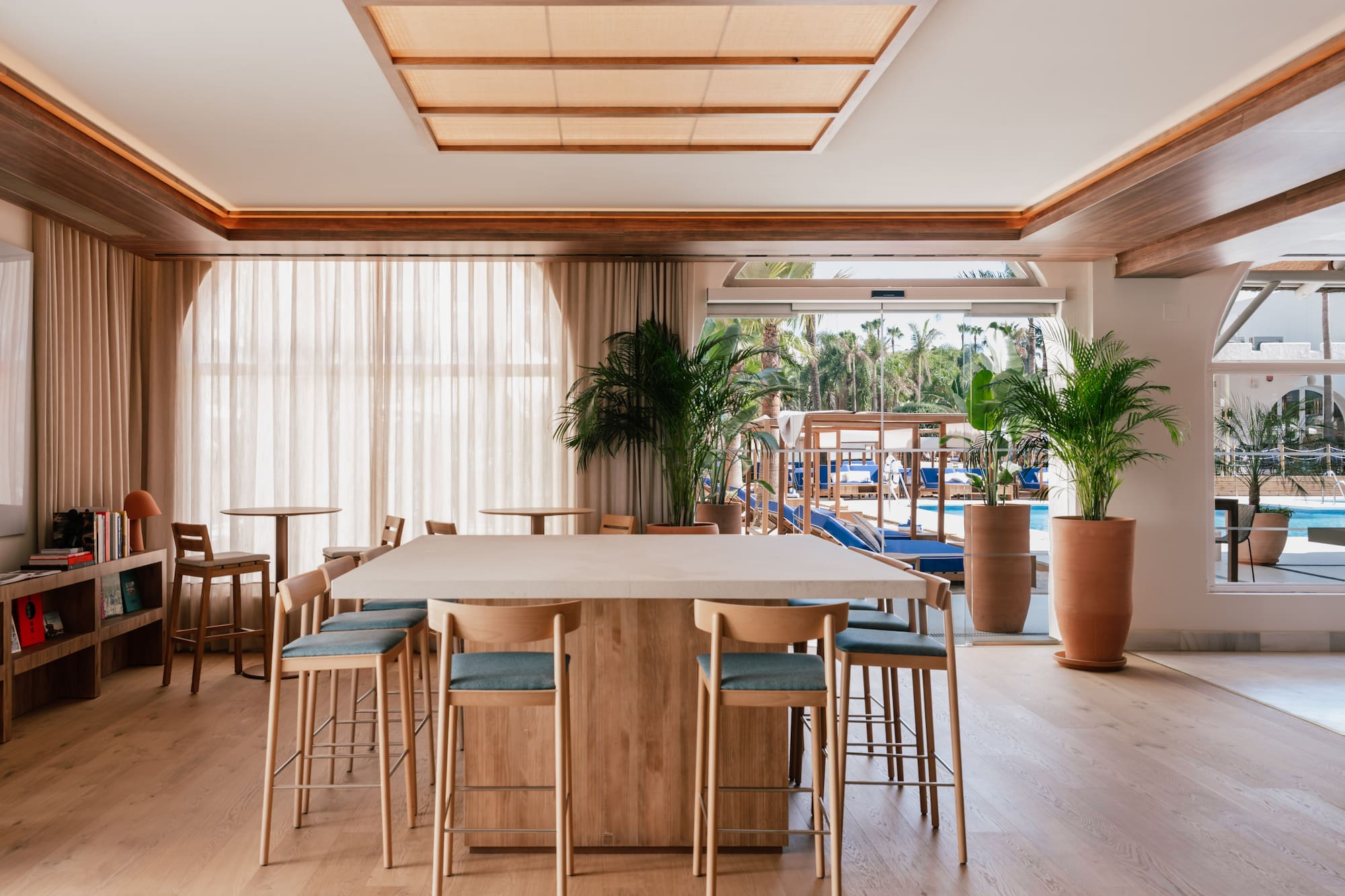 Hotel communal lounge with bar table, wooden stools, plants, and pool view.