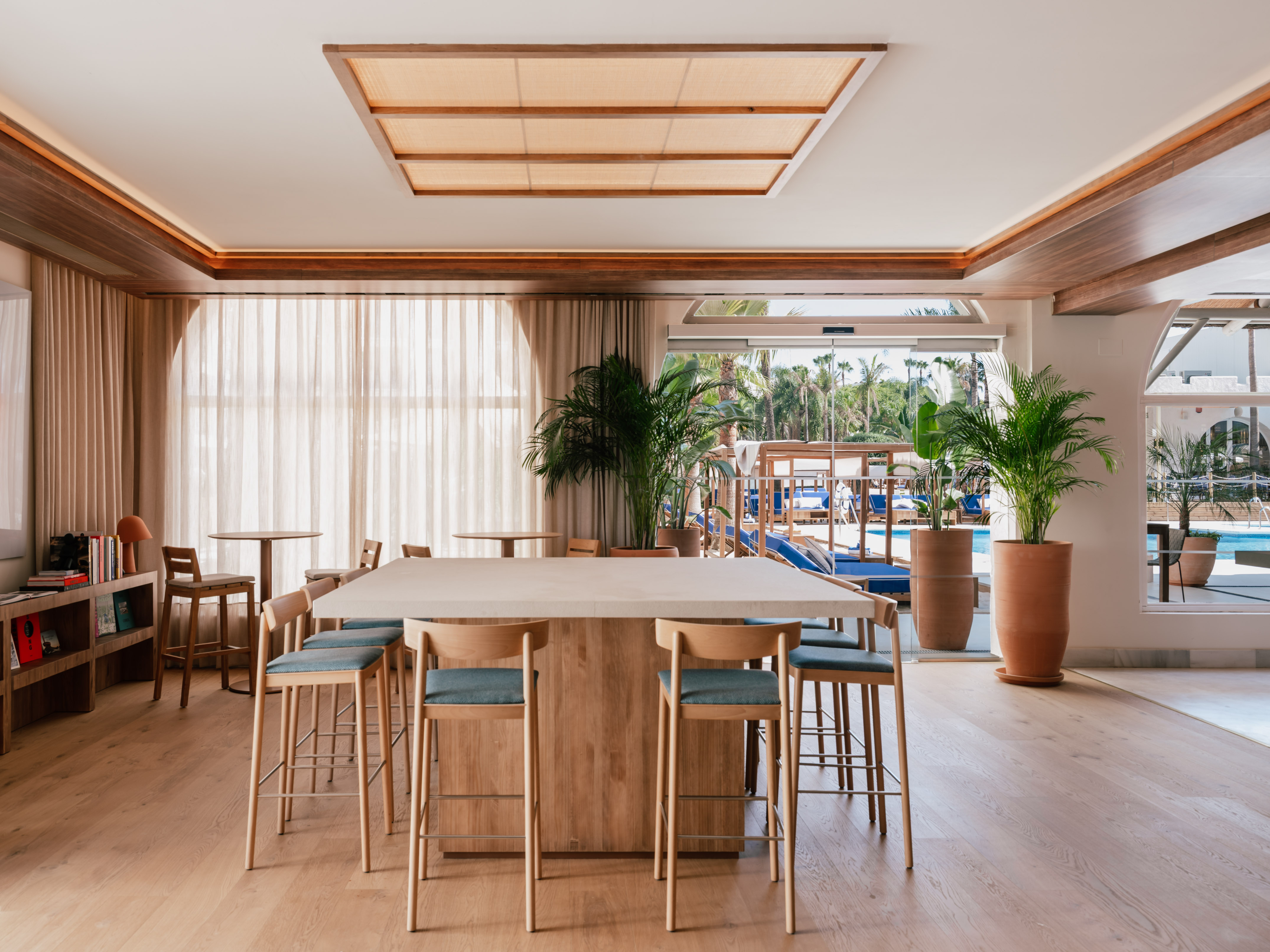 Hotel communal lounge with bar table, wooden stools, plants, and pool view.