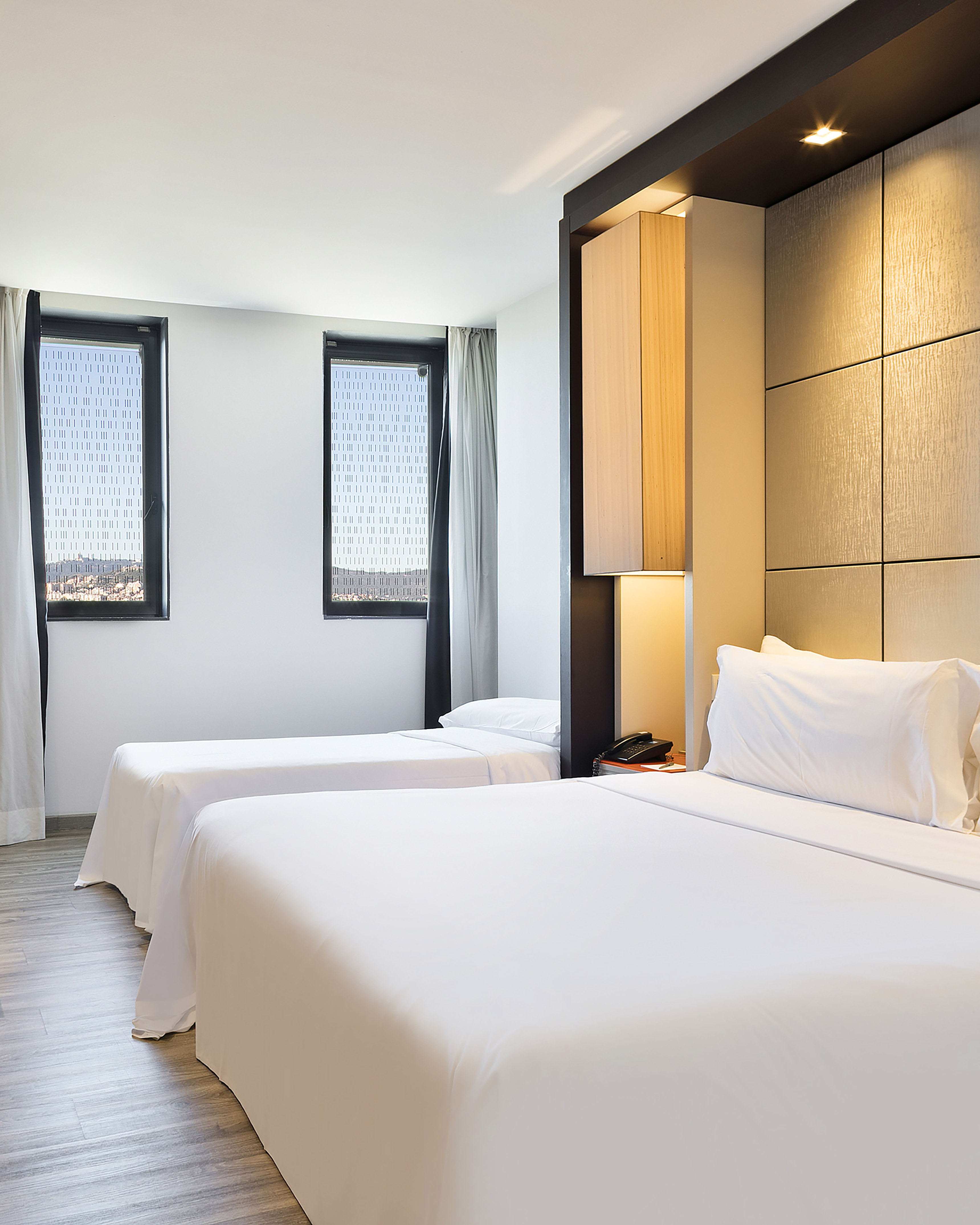Bright hotel room with two white beds, modern headboard, and city view windows.