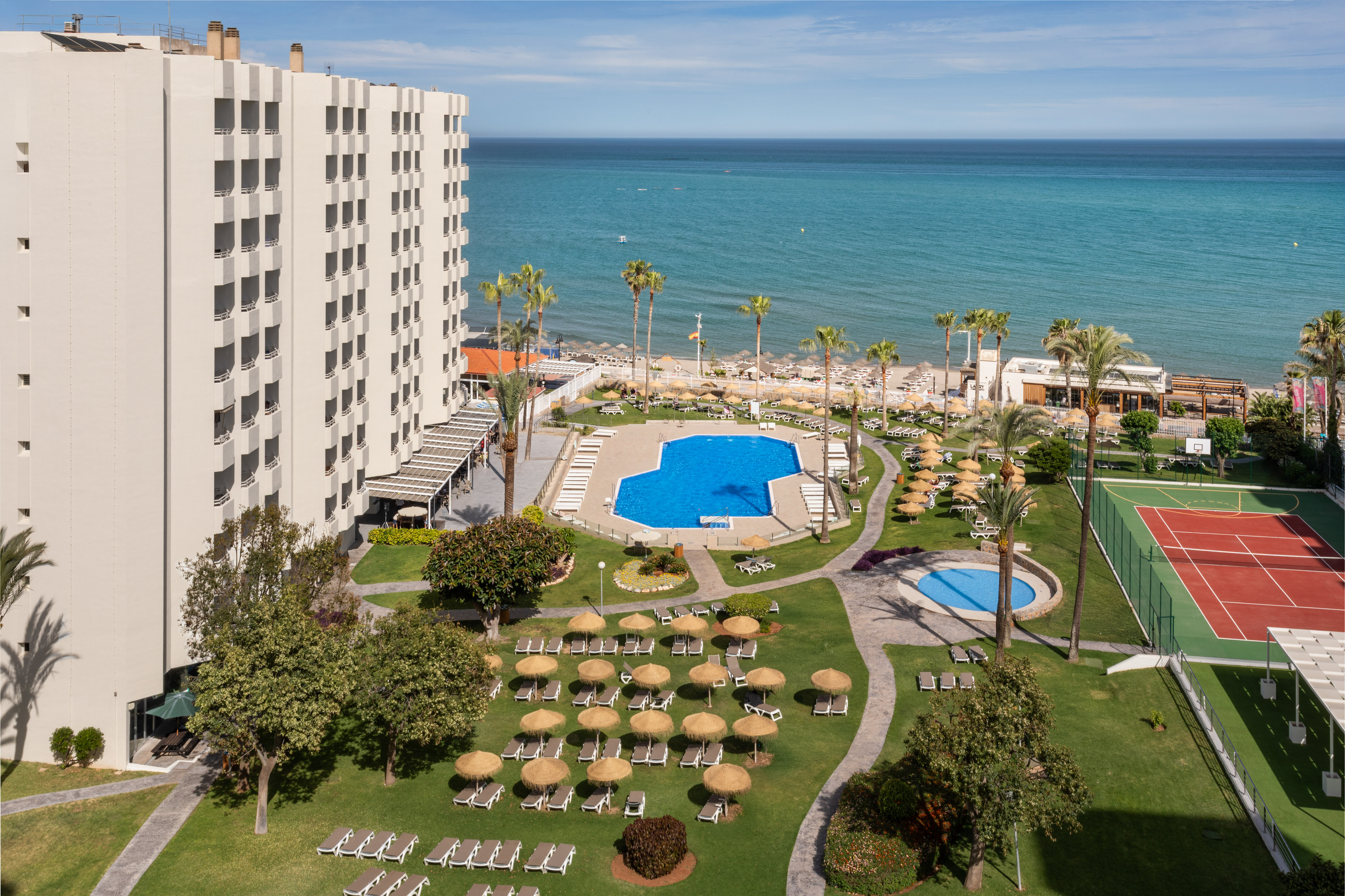 Oceanfront hotel resort with large pool, sandy beach, palm trees, and tennis court.