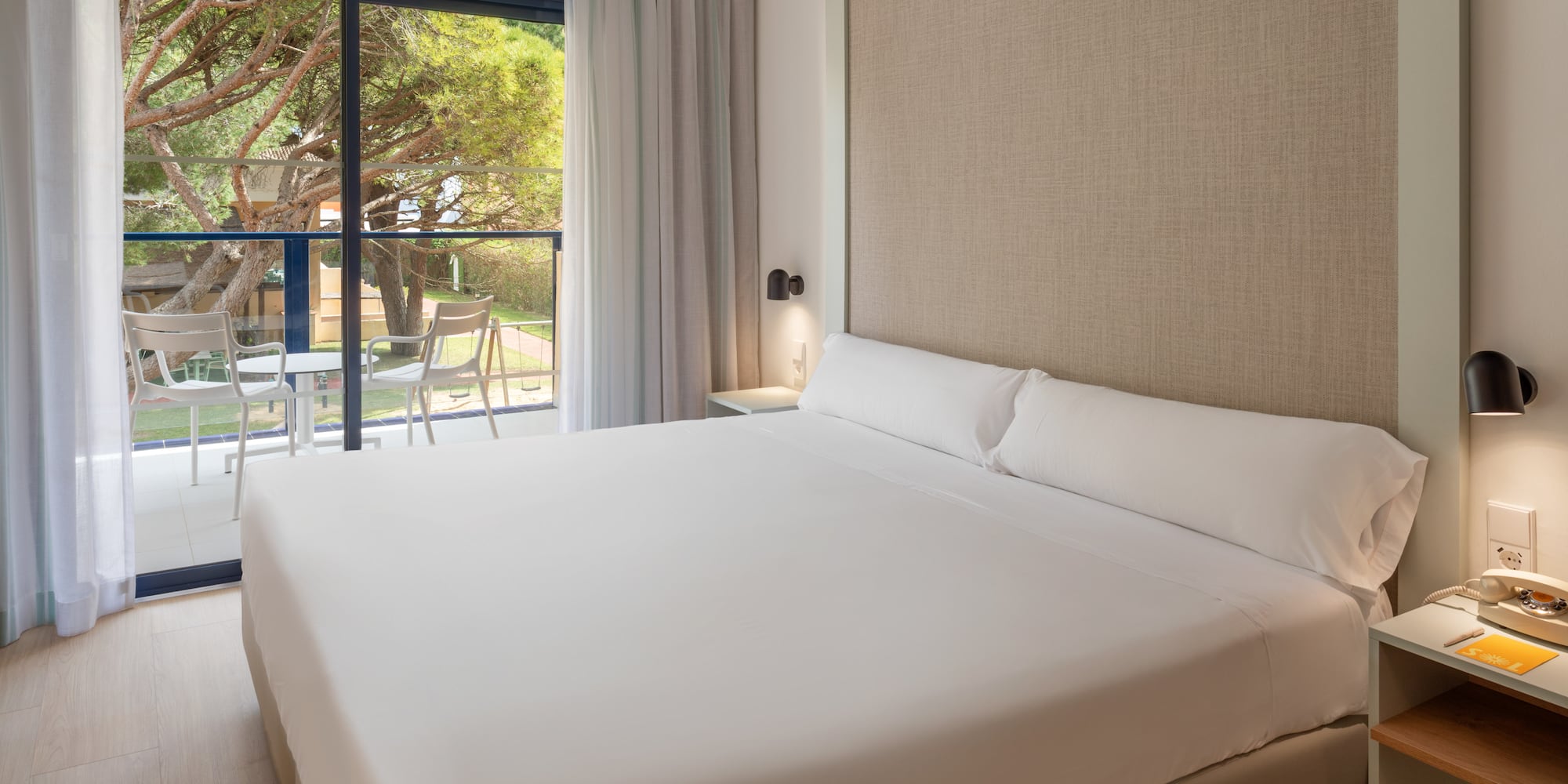 Modern hotel room with white bed, balcony, and natural tree landscape.