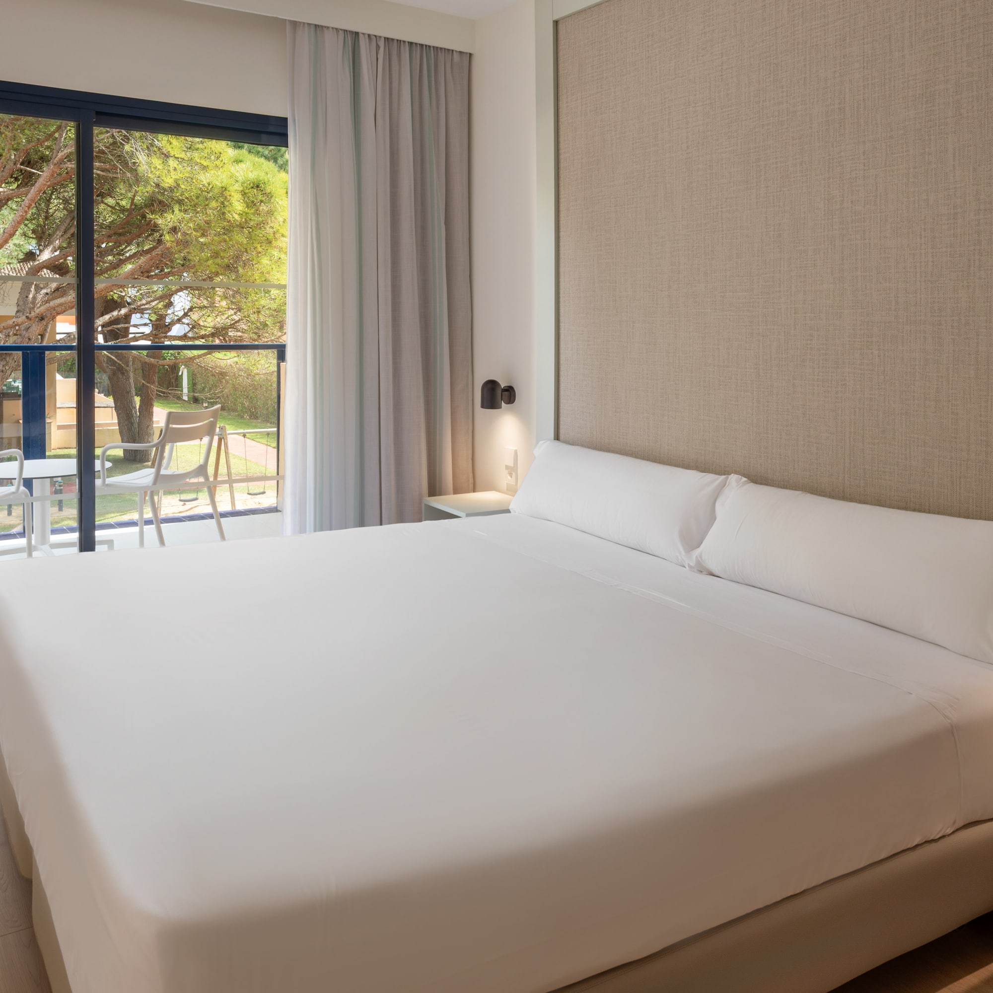 Modern hotel room with white bed, balcony, and natural tree landscape.