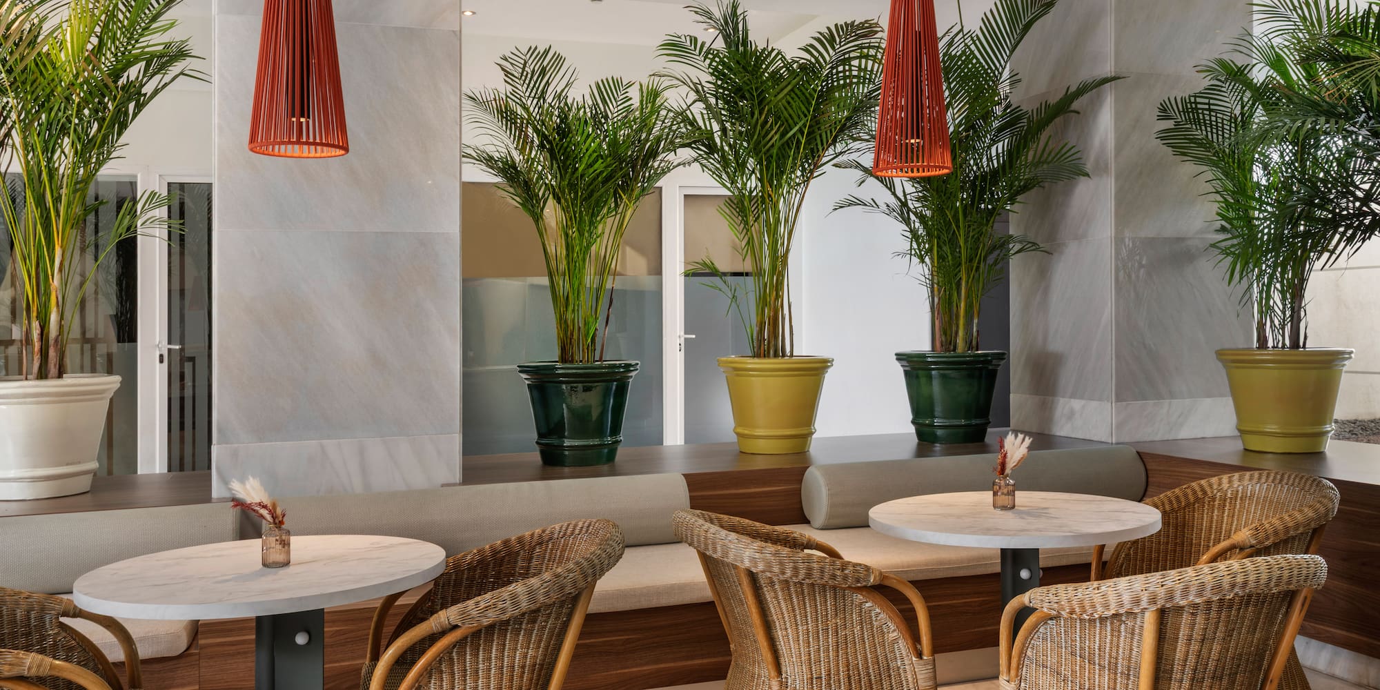 Modern hotel lounge with wicker chairs, marble tables, and potted palm trees.