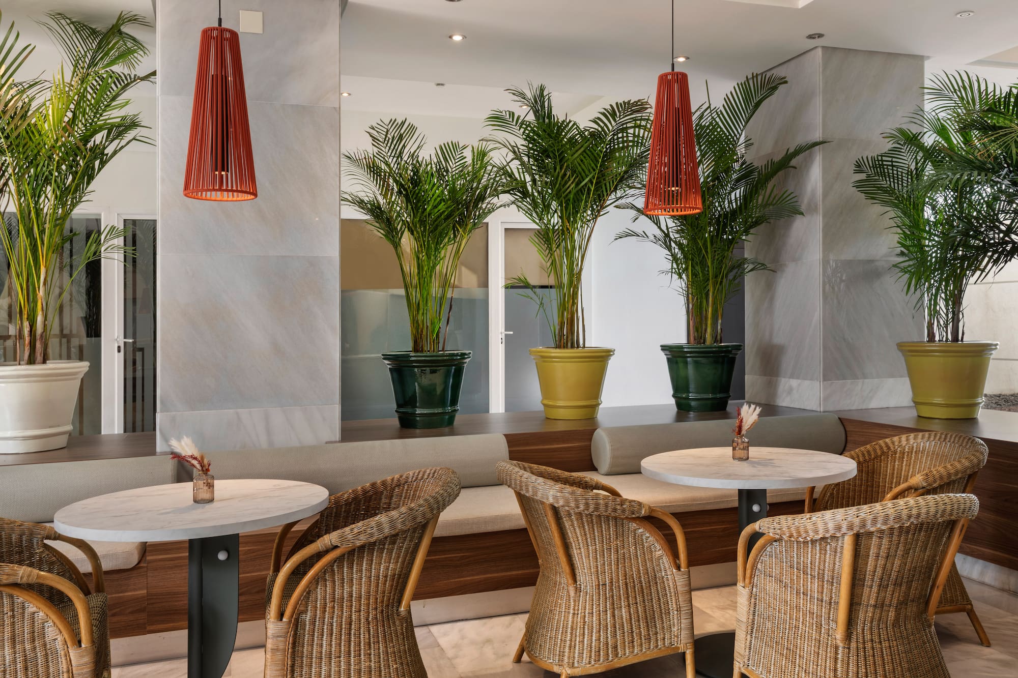 Modern hotel lounge with wicker chairs, marble tables, and potted palm trees.
