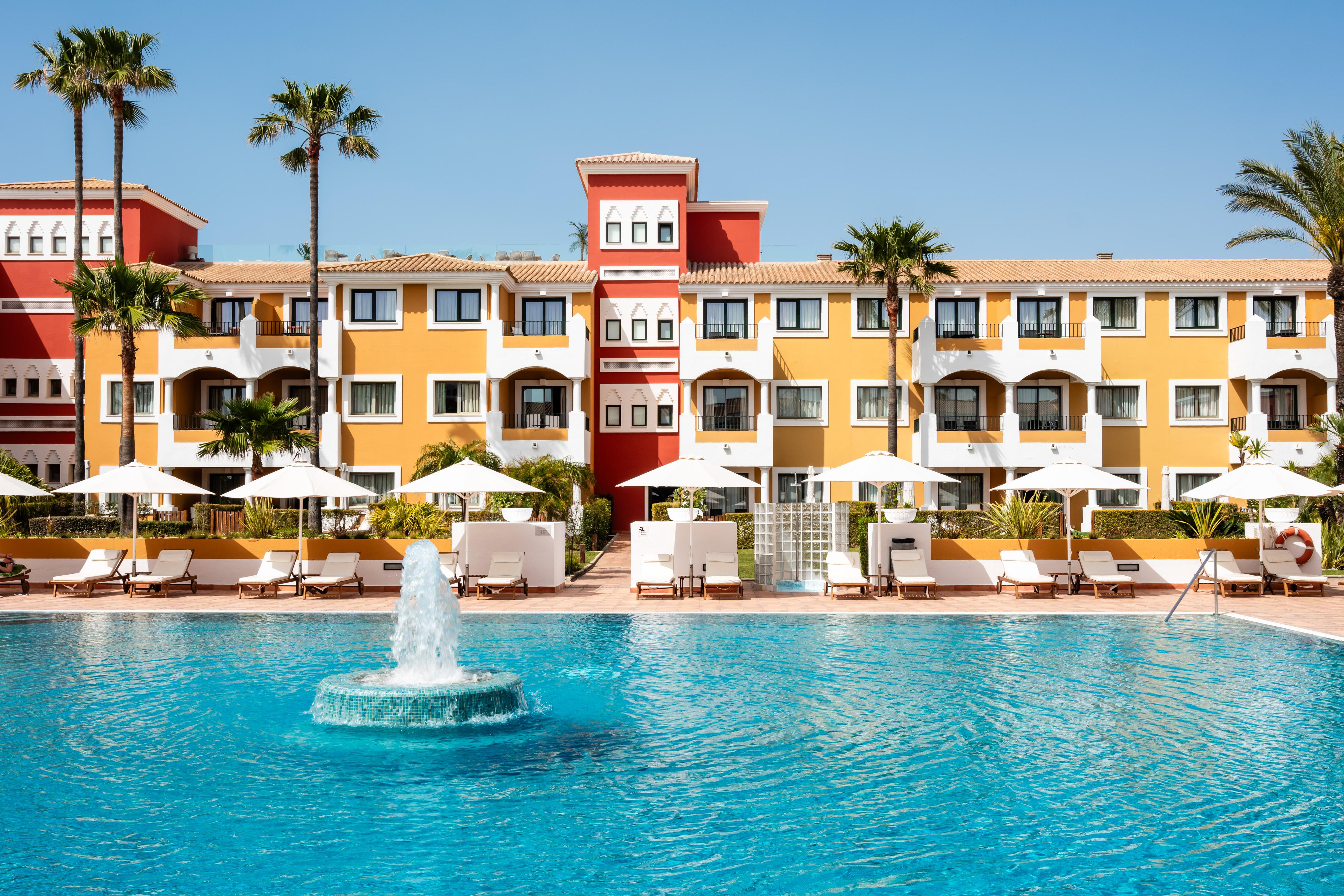 Yellow and red hotel building, palm trees, large pool with fountain.