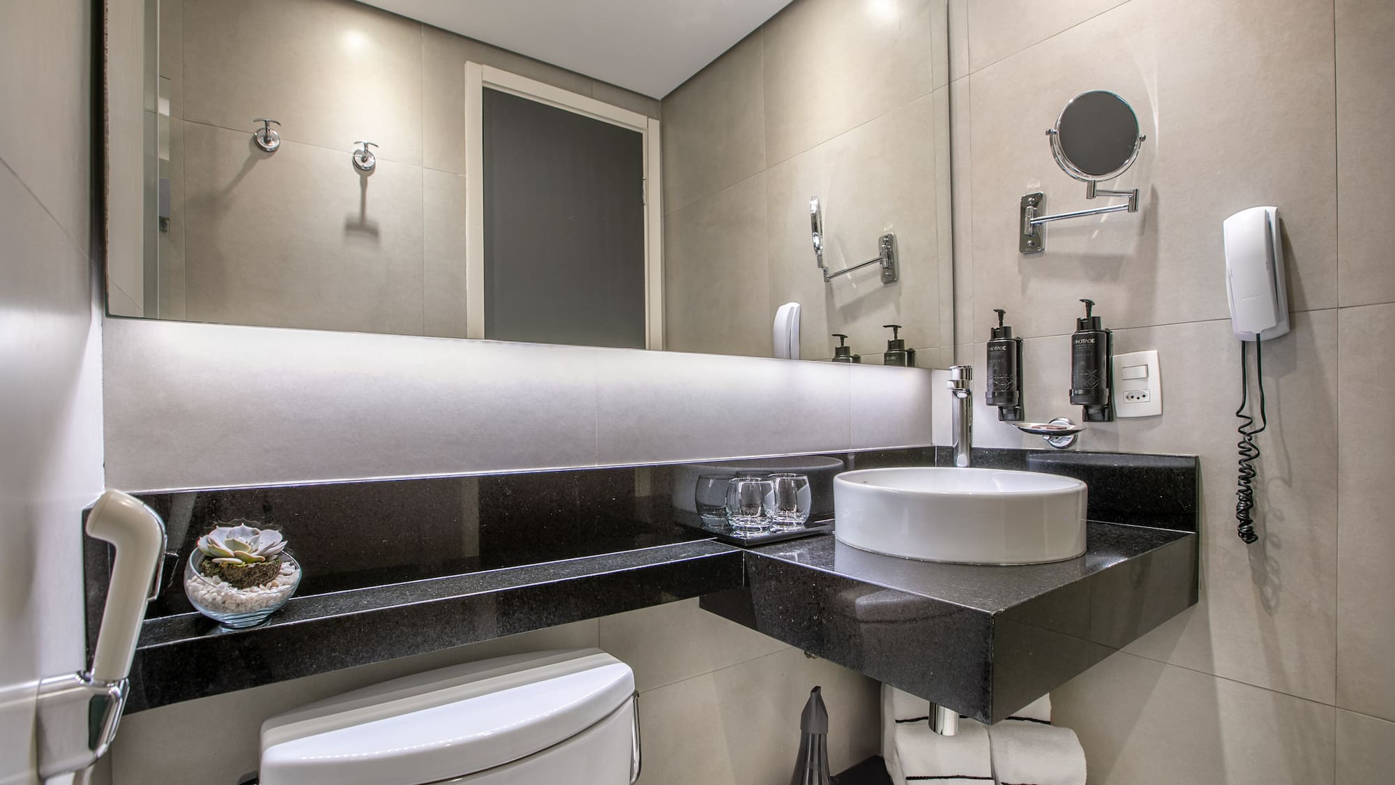 Banheiro moderno com pia branca, bancada preta e espelho iluminado. - Deluxe Room Queen - Meliá Ibirapuera, São Paulo