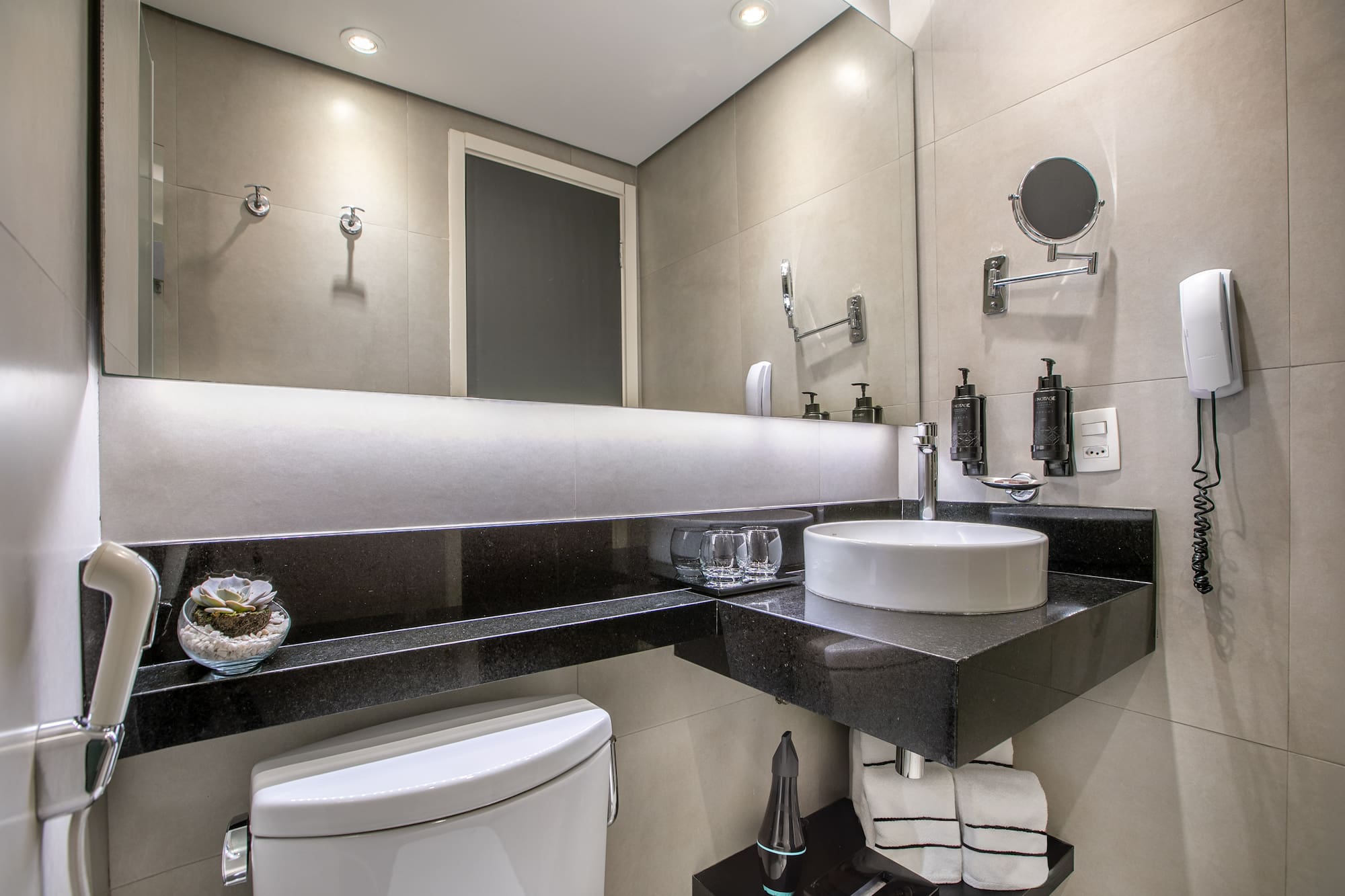 Banheiro moderno com pia branca, bancada preta e espelho iluminado. - Deluxe Room Queen - Meliá Ibirapuera, São Paulo