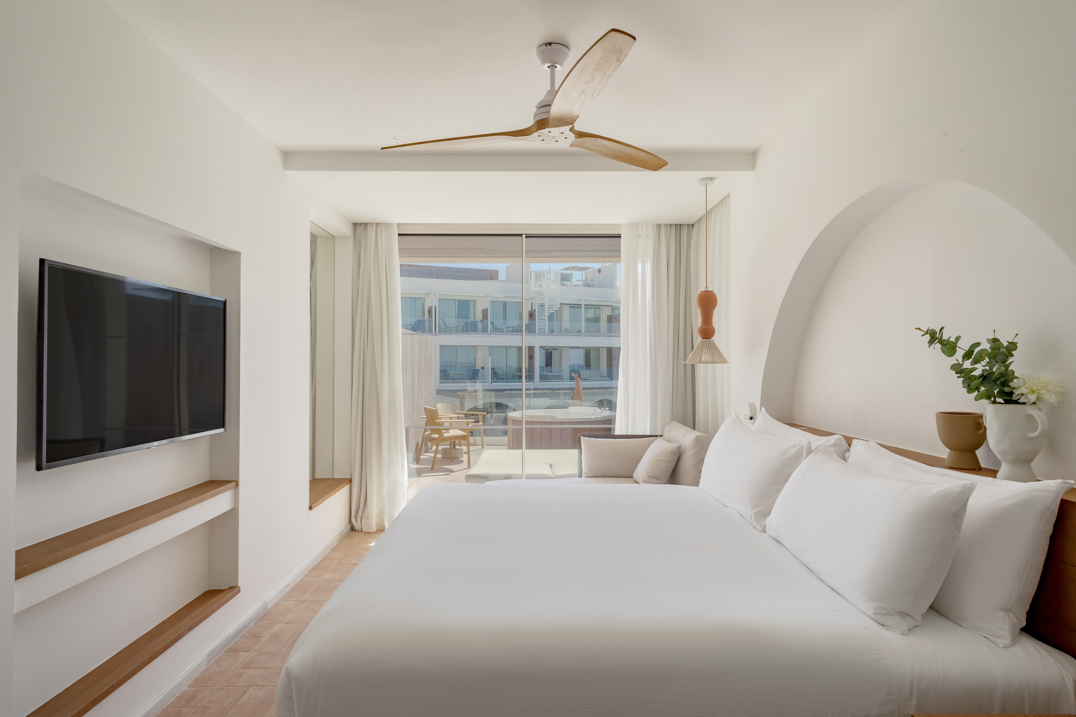 Modern resort bedroom with white bed, balcony, and wooden ceiling fan.