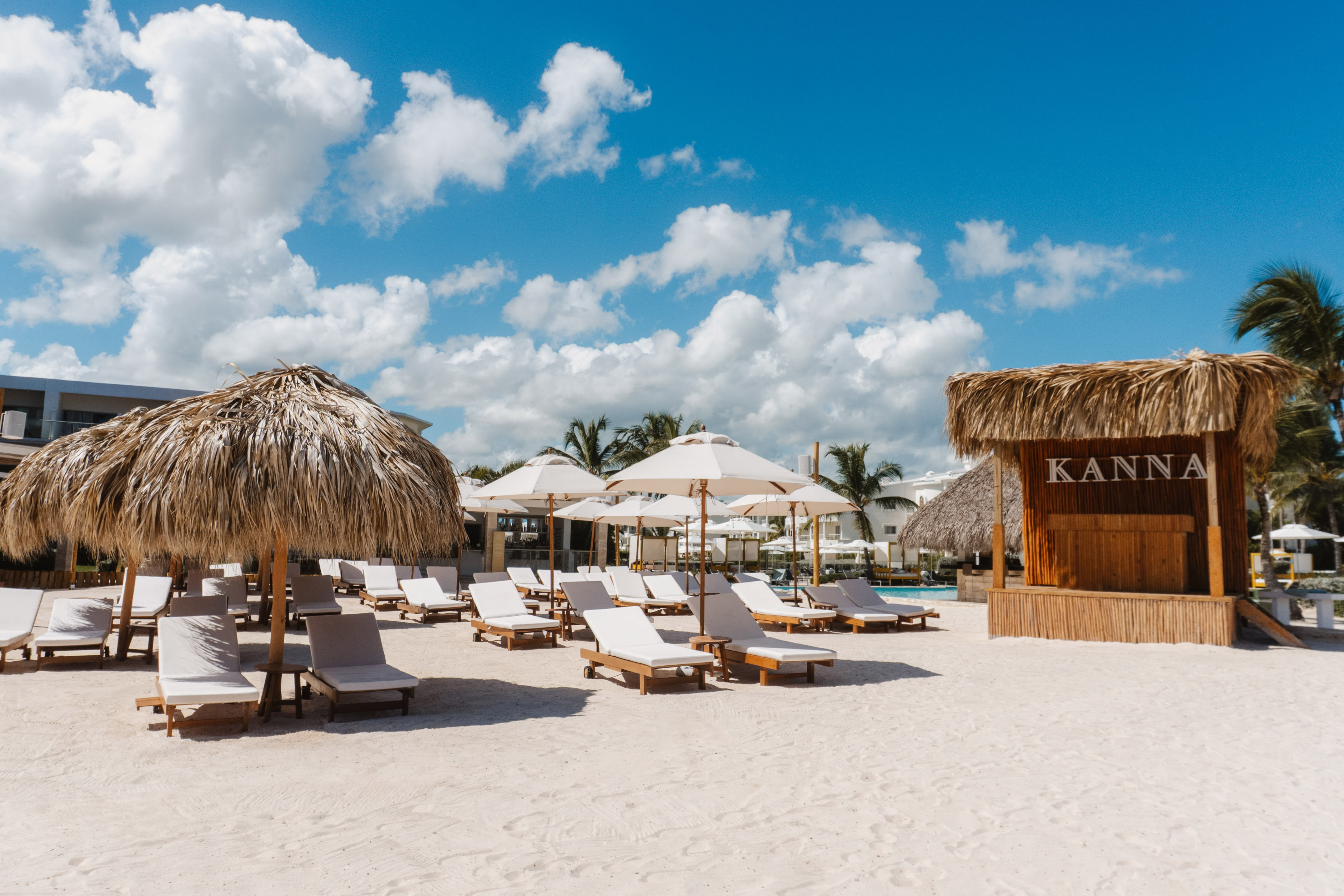 Sunny resort beach with KANNA bar, white lounge chairs, and umbrellas.