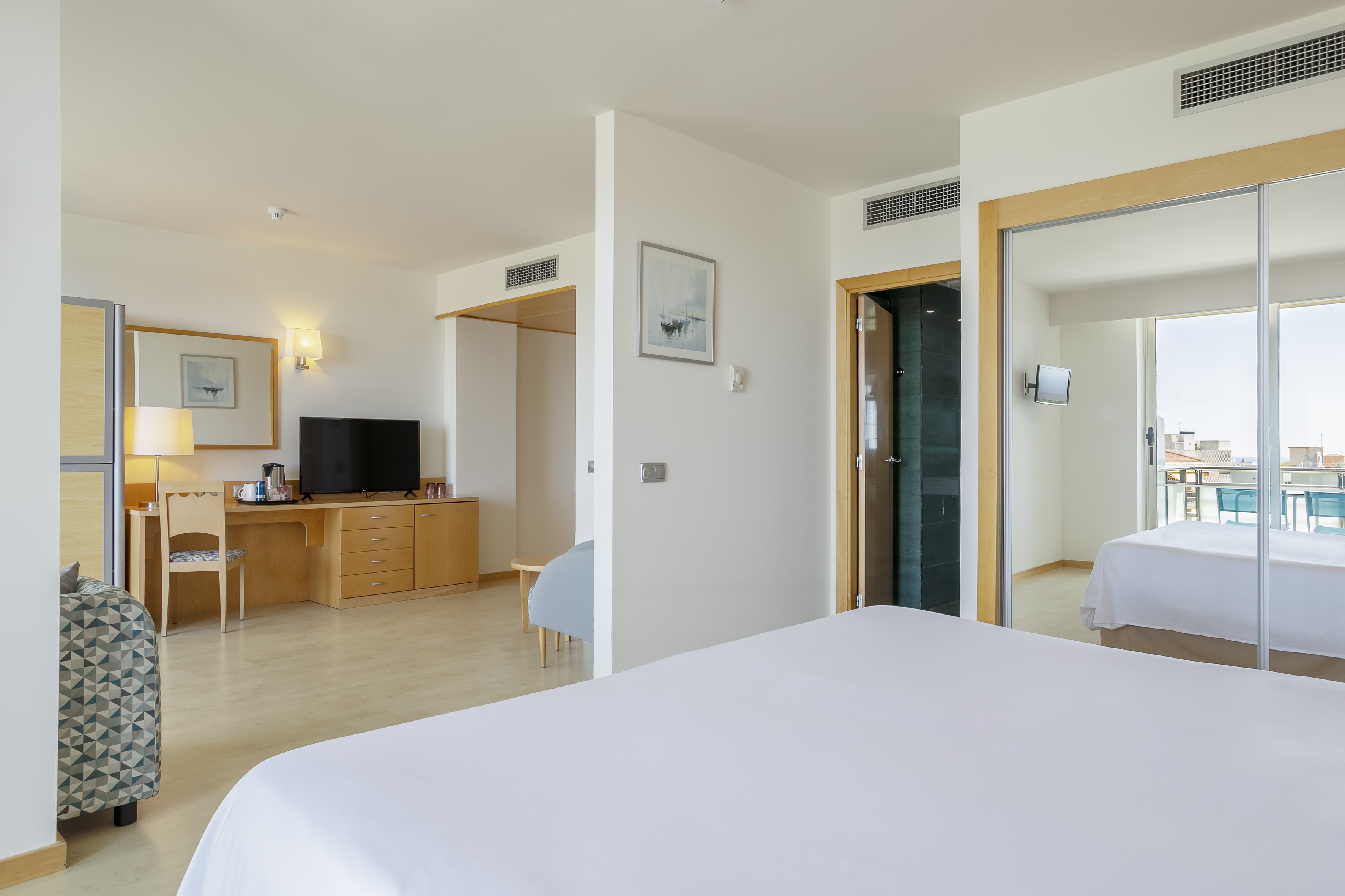 Modern hotel suite with bed, TV, desk, and bright balcony view.