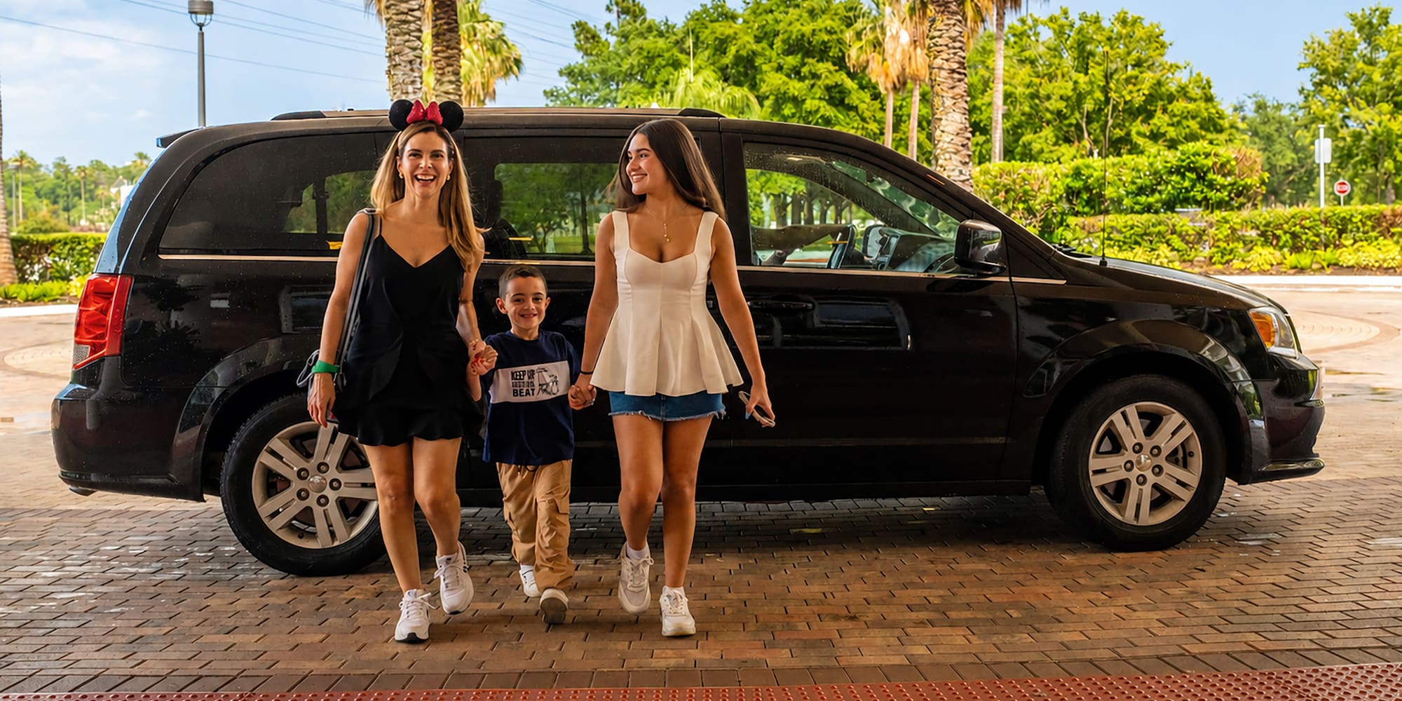 Family (Minnie ears) walks from black minivan at sunny resort.