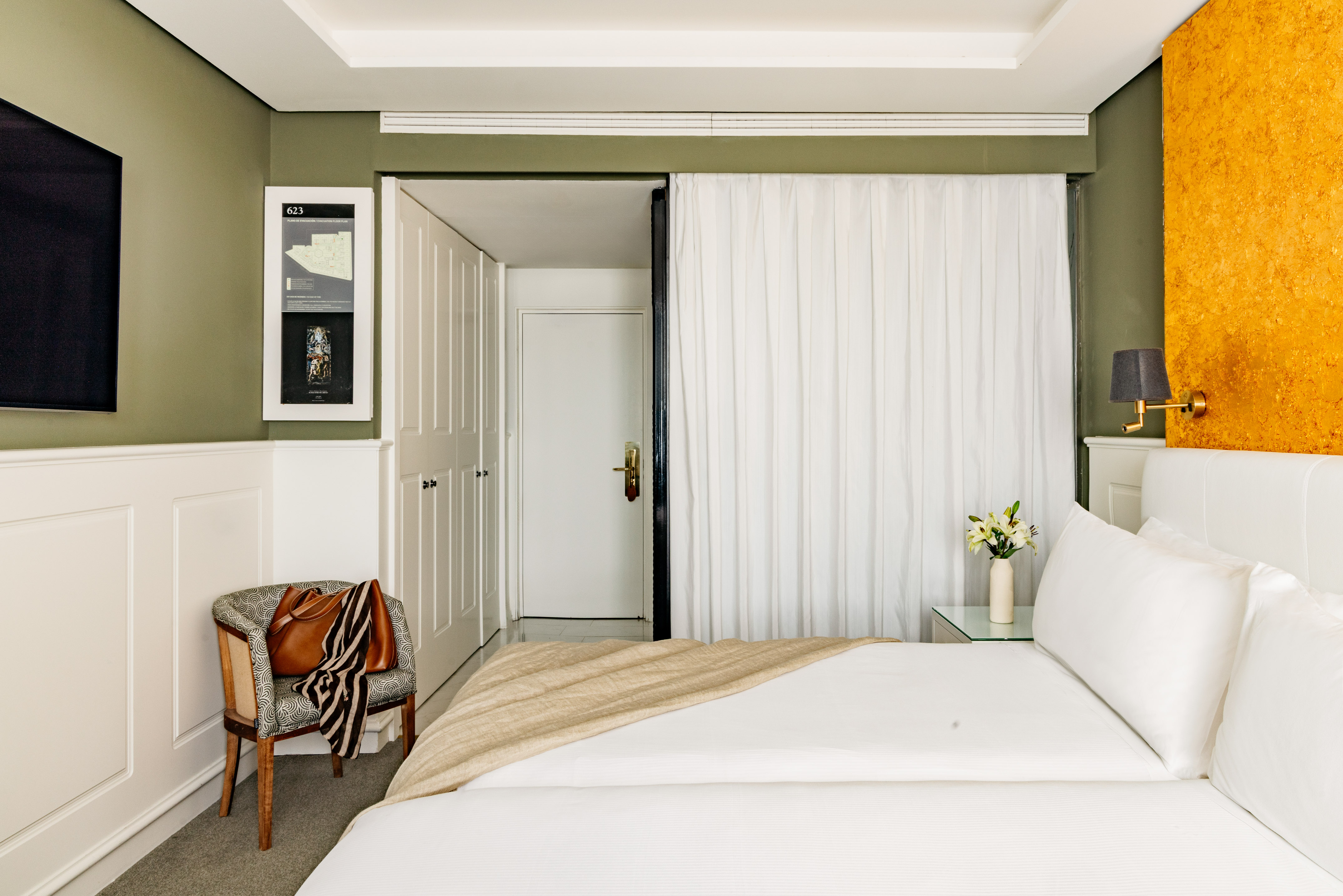 Stylish hotel room with white bed, olive green walls, and vibrant orange accent wall.