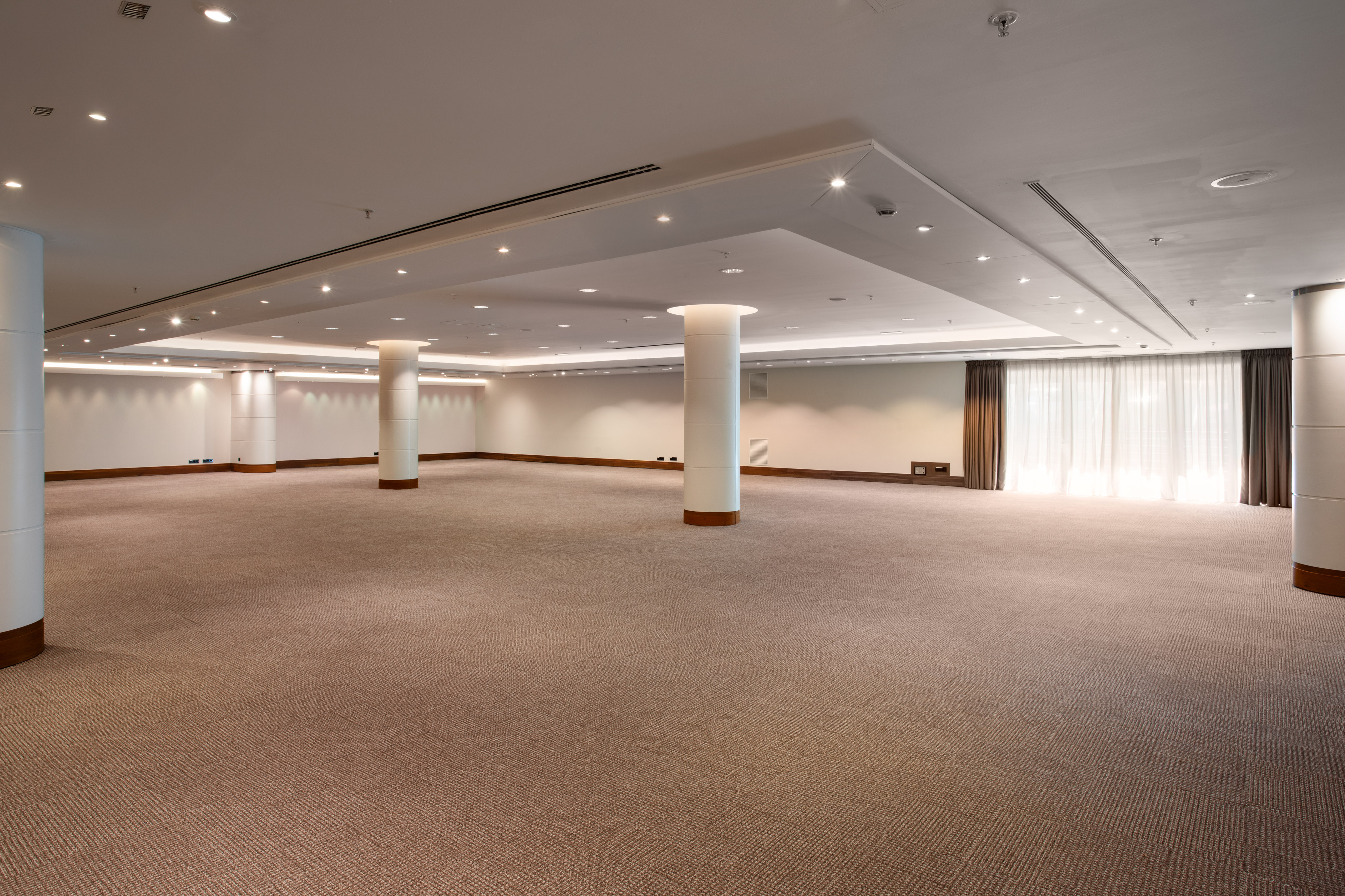 Modern empty event space with pillars, carpeted floor, and bright curtains.