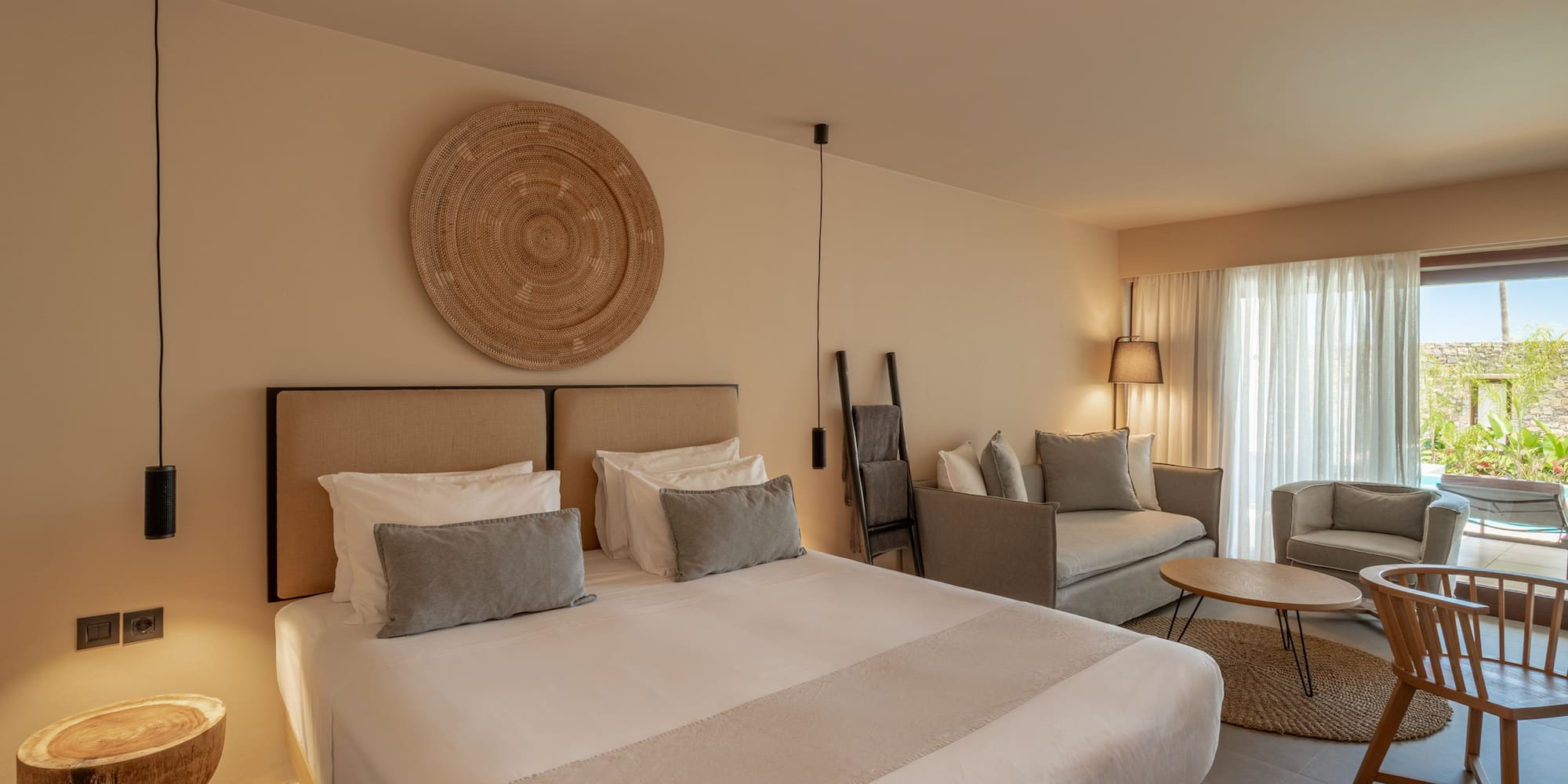 Neutral modern hotel suite with bed, woven wall decor, and a living area.