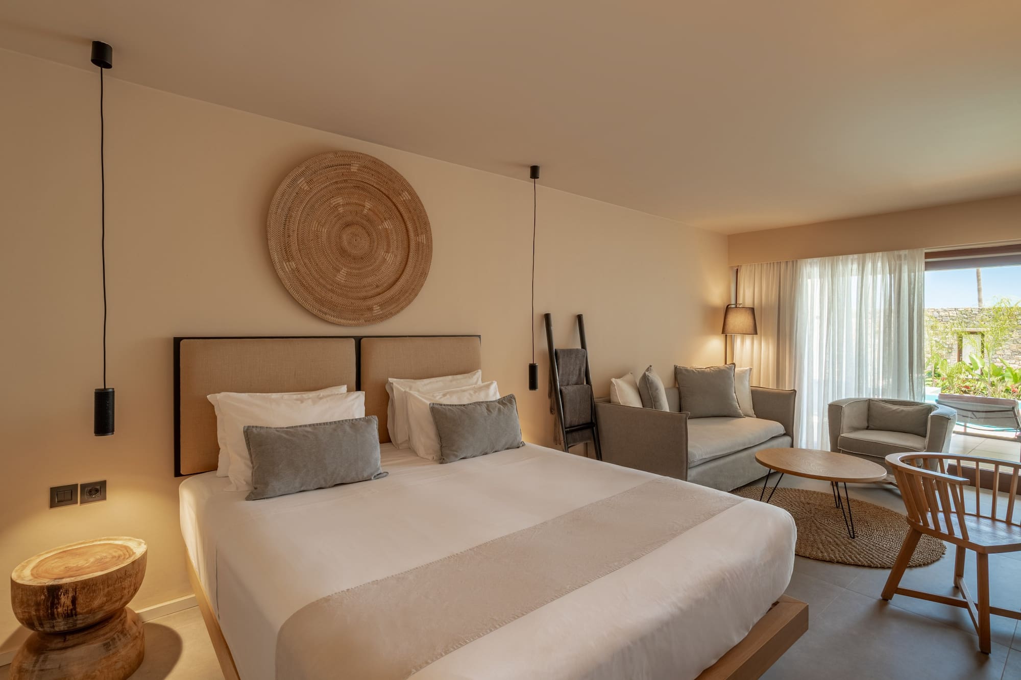 Neutral modern hotel suite with bed, woven wall decor, and a living area.