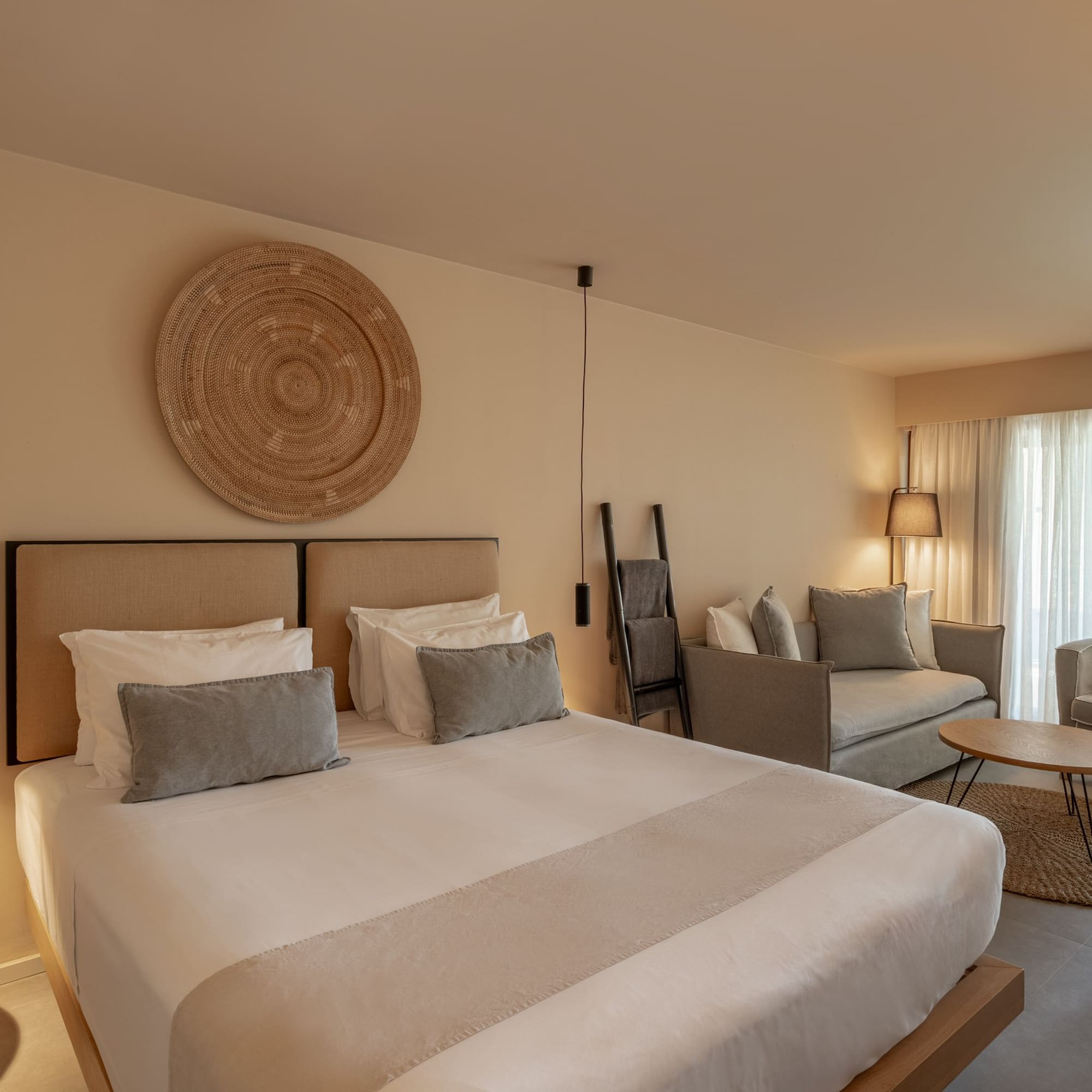 Neutral modern hotel suite with bed, woven wall decor, and a living area.