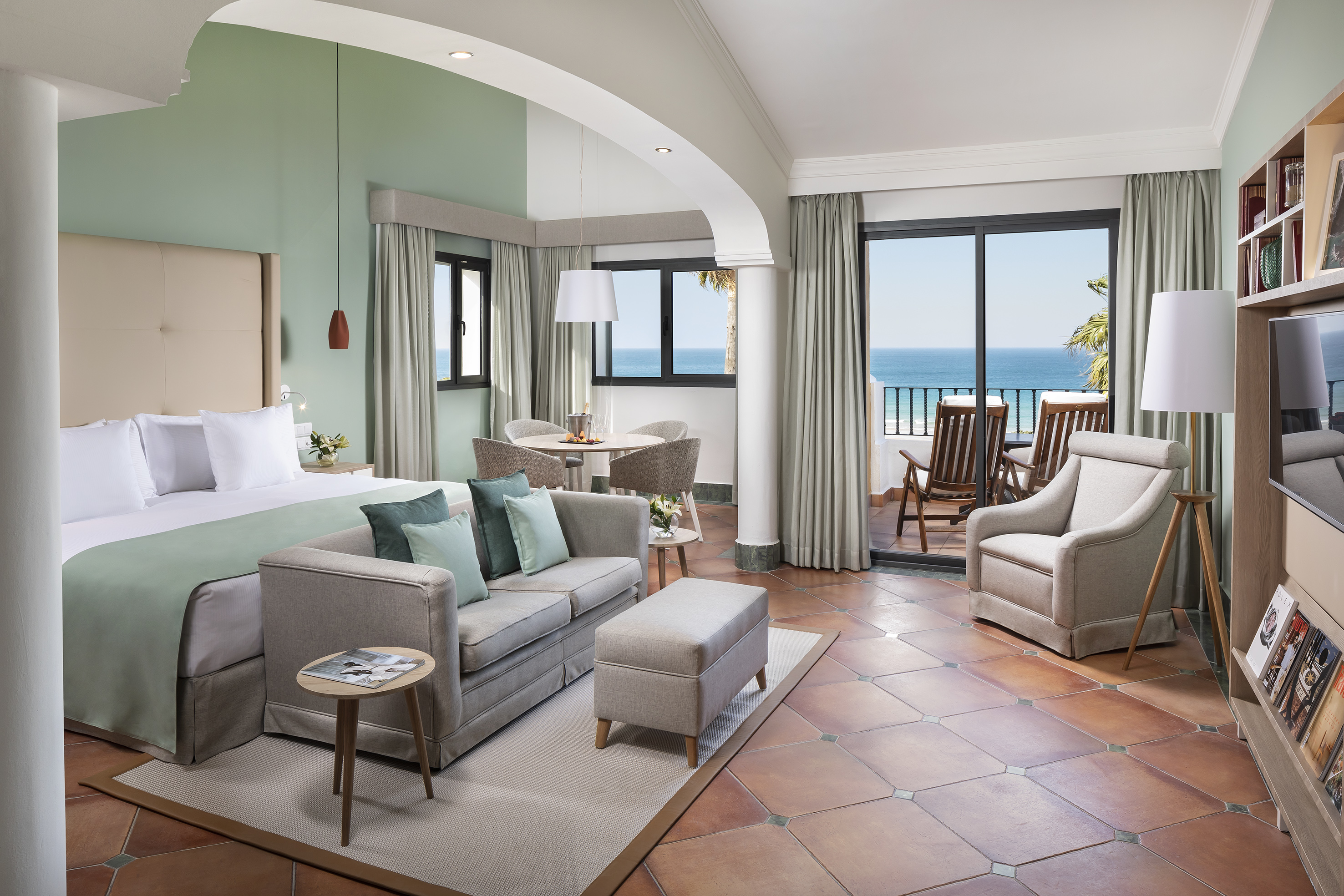 Modern hotel suite with ocean view balcony, bed, sofa, and dining area.