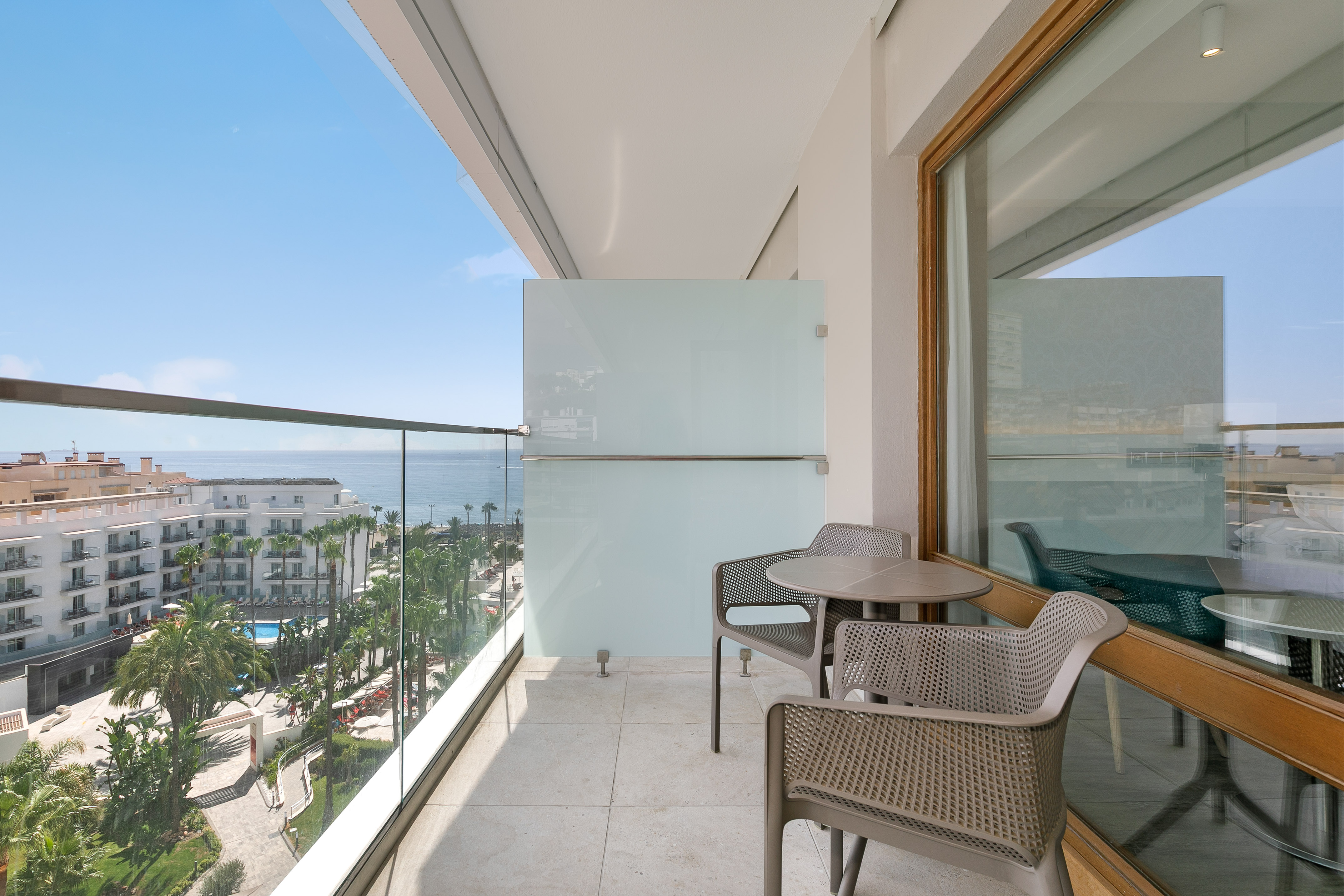 Balcony with seating, glass railing, and sea view overlooking a resort.