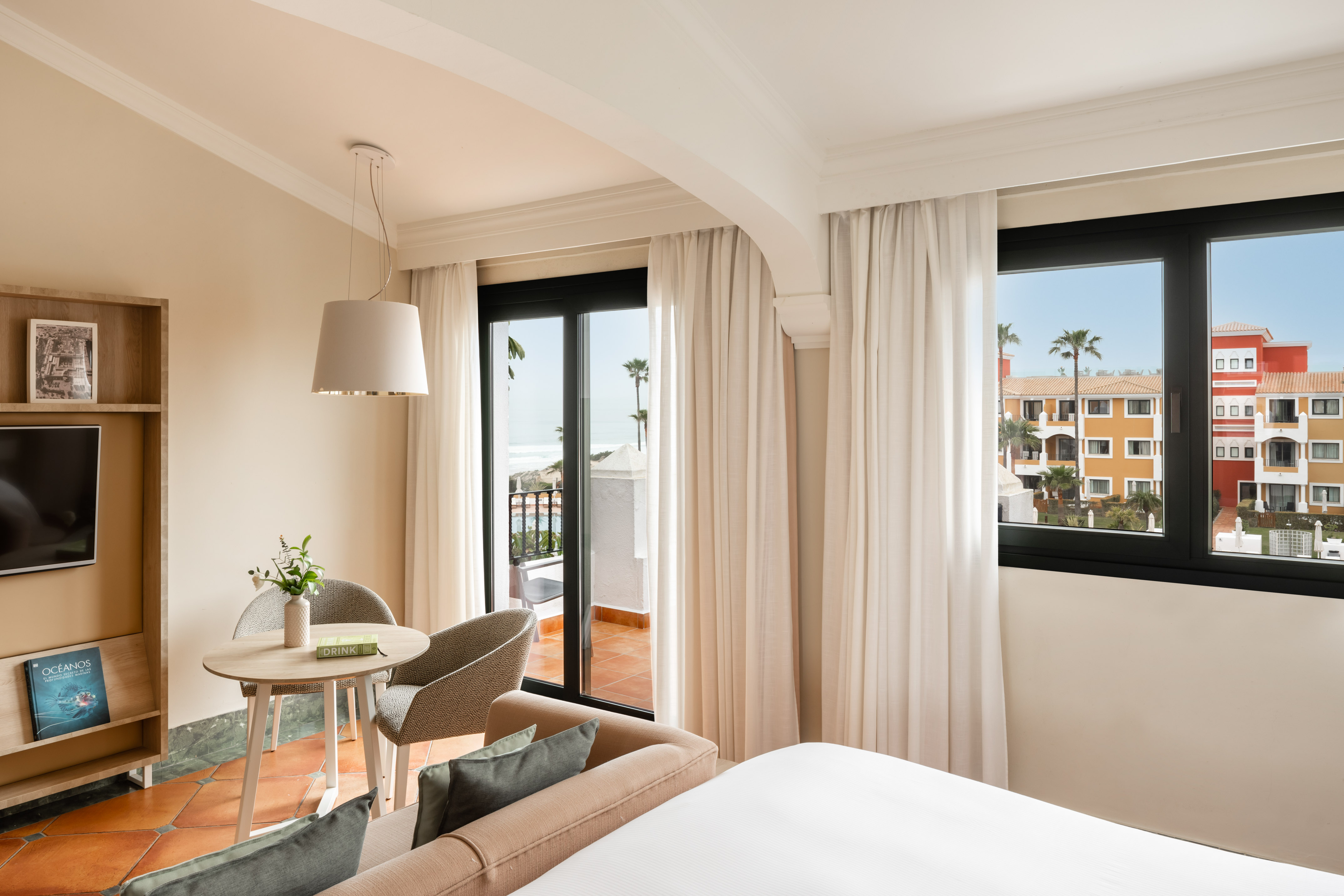Ocean view hotel room with balcony, palm trees, and resort buildings.
