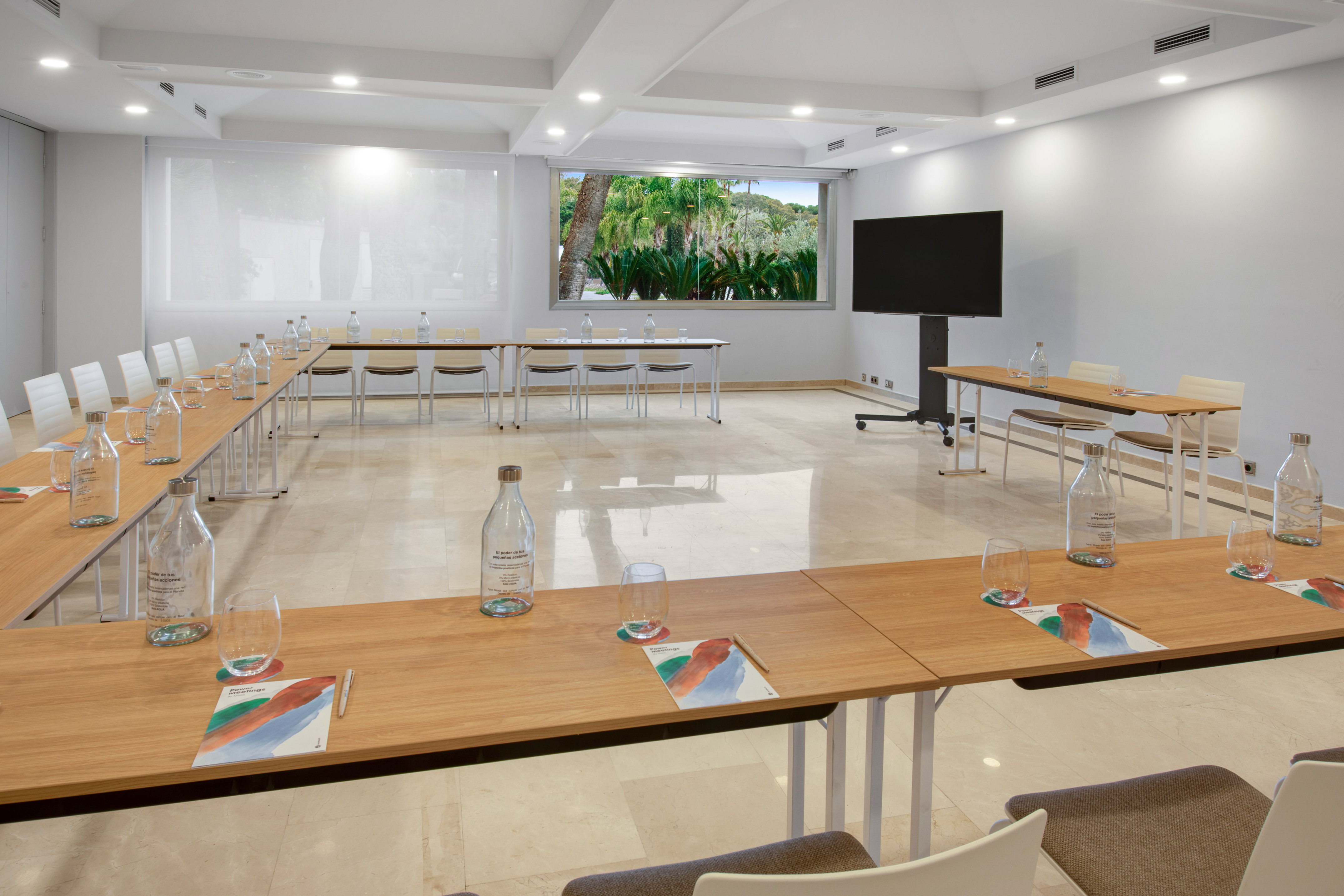 Modern meeting room with U-shaped table, tropical view, and TV screen.
