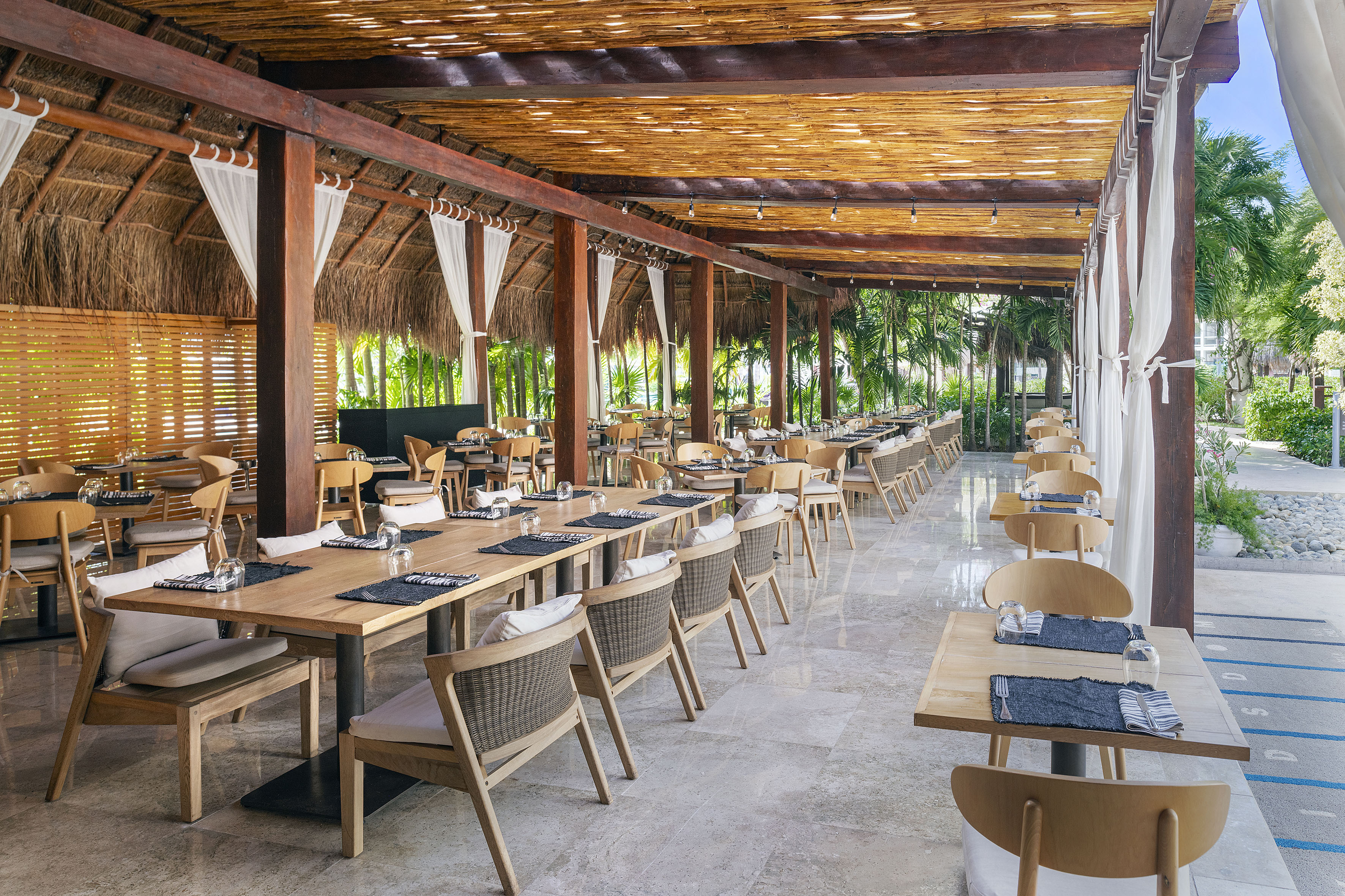 Tropical outdoor restaurant with palapa roof, wooden tables, and chairs.