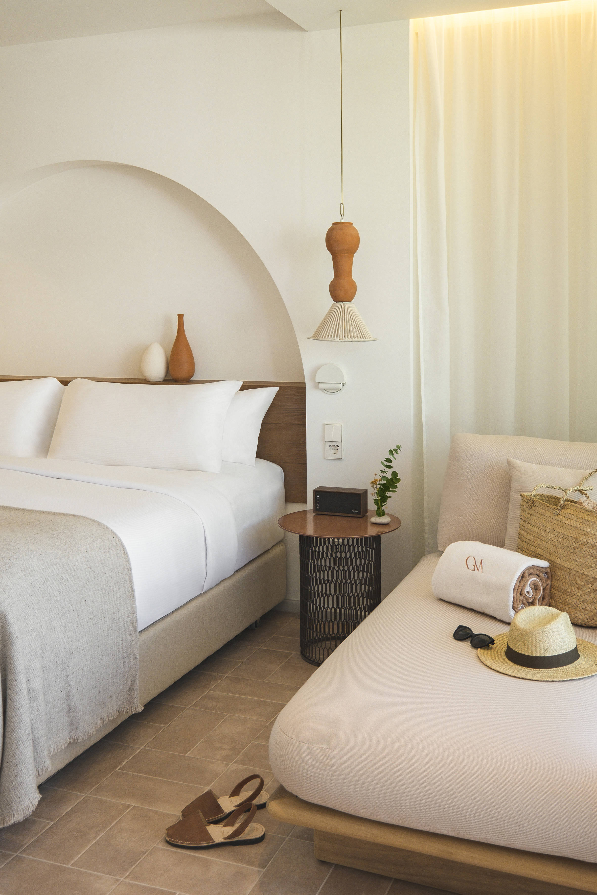 Modern hotel room with white bed, lounge chair, straw hat, and terracotta decor.