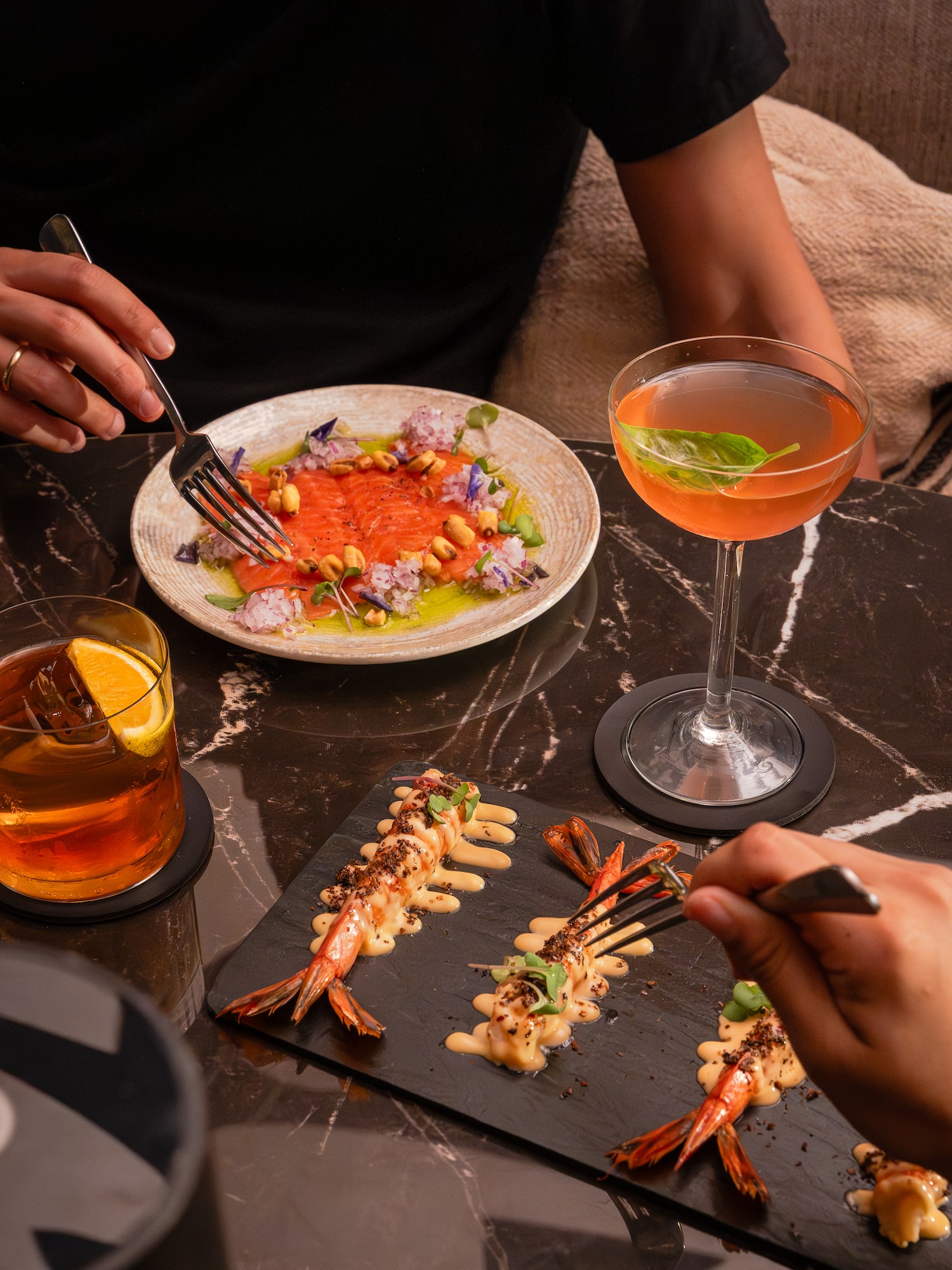 Cena con carpaccio de salmón, langostinos y cócteles en mesa de mármol oscuro.