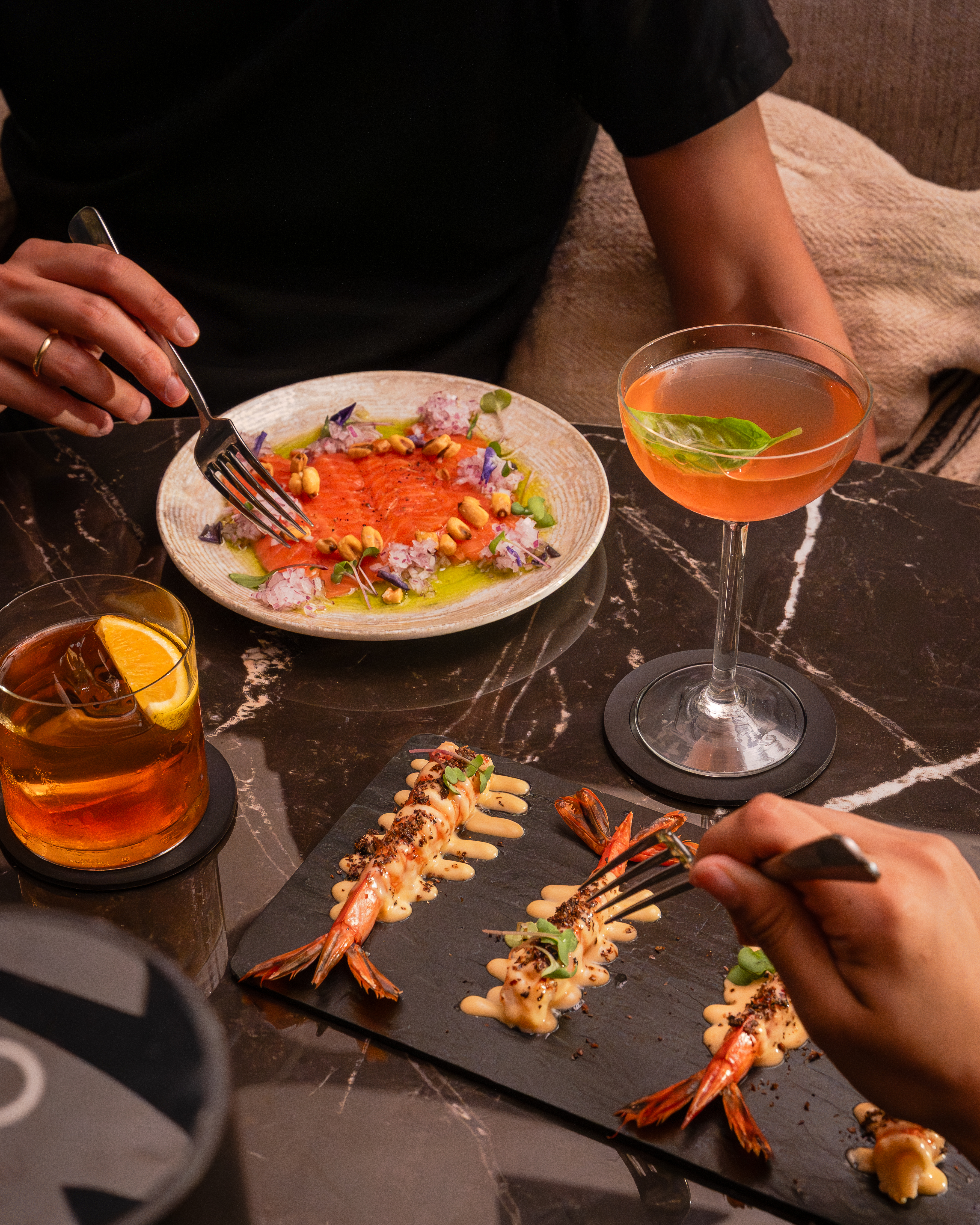 Cena con carpaccio de salmón, langostinos y cócteles en mesa de mármol oscuro.