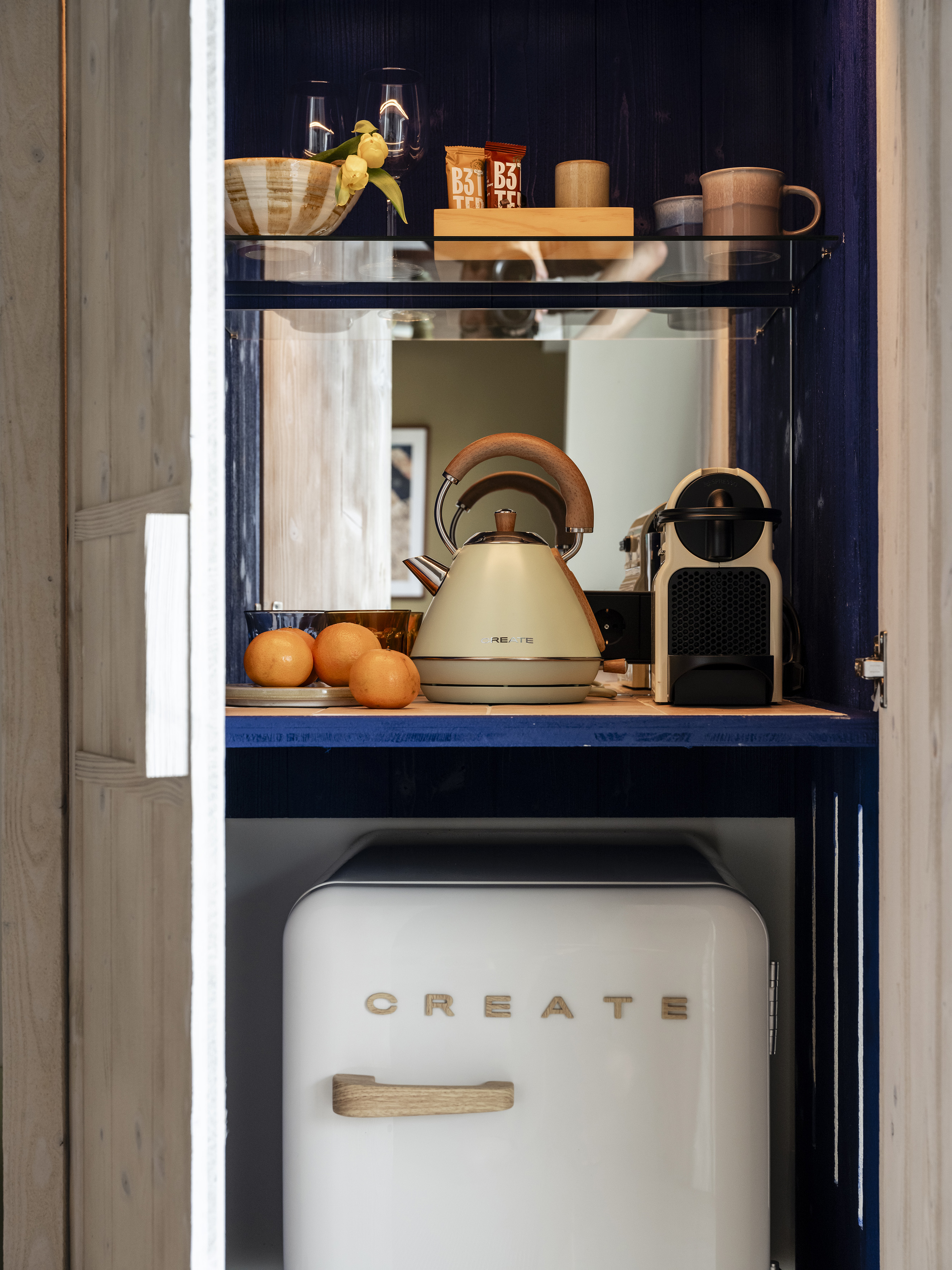 Hotel minibar with coffee maker, cream kettle, snacks, and white retro fridge.