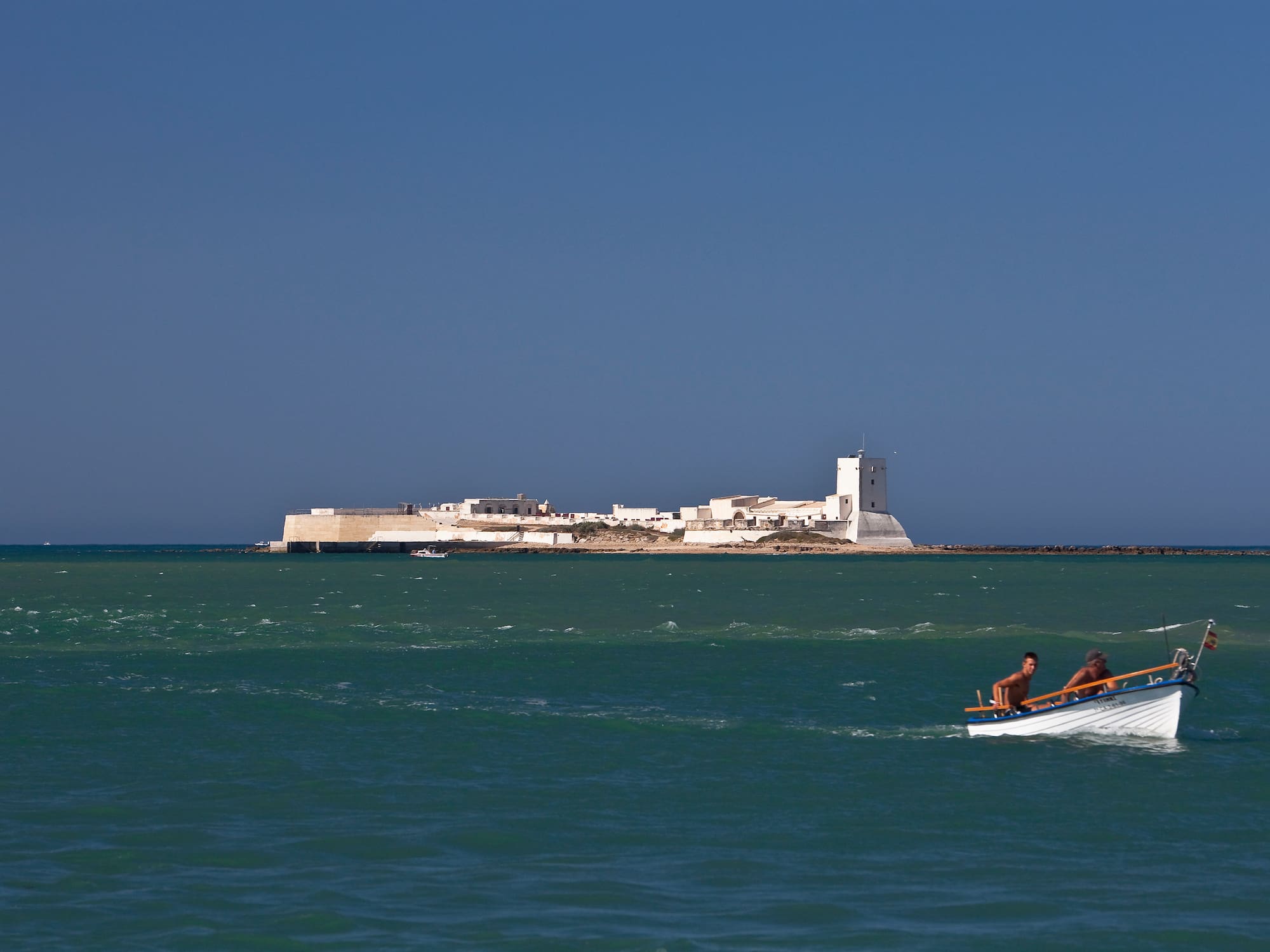Men in boat on sea, white San Sebastian Castle fortress in Cadiz, Spain.