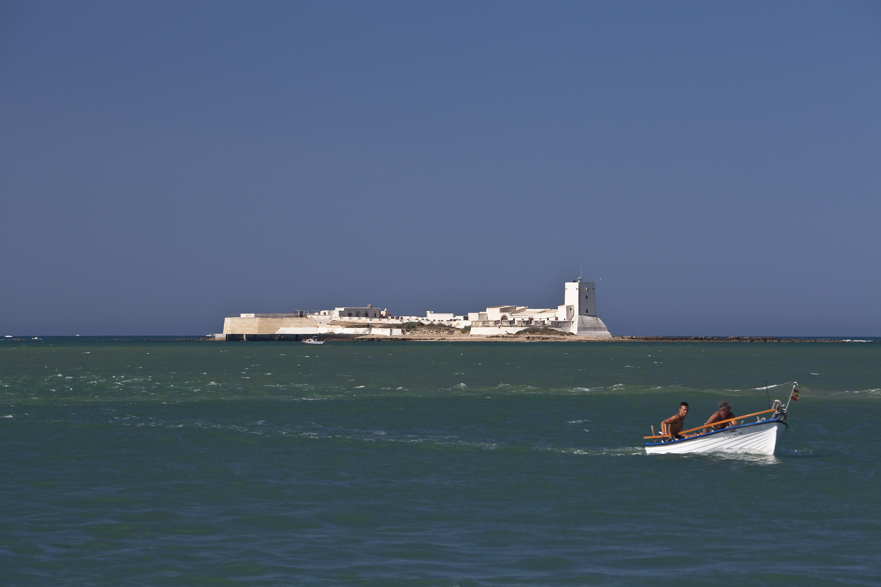 Men in boat on sea, white San Sebastian Castle fortress in Cadiz, Spain.
