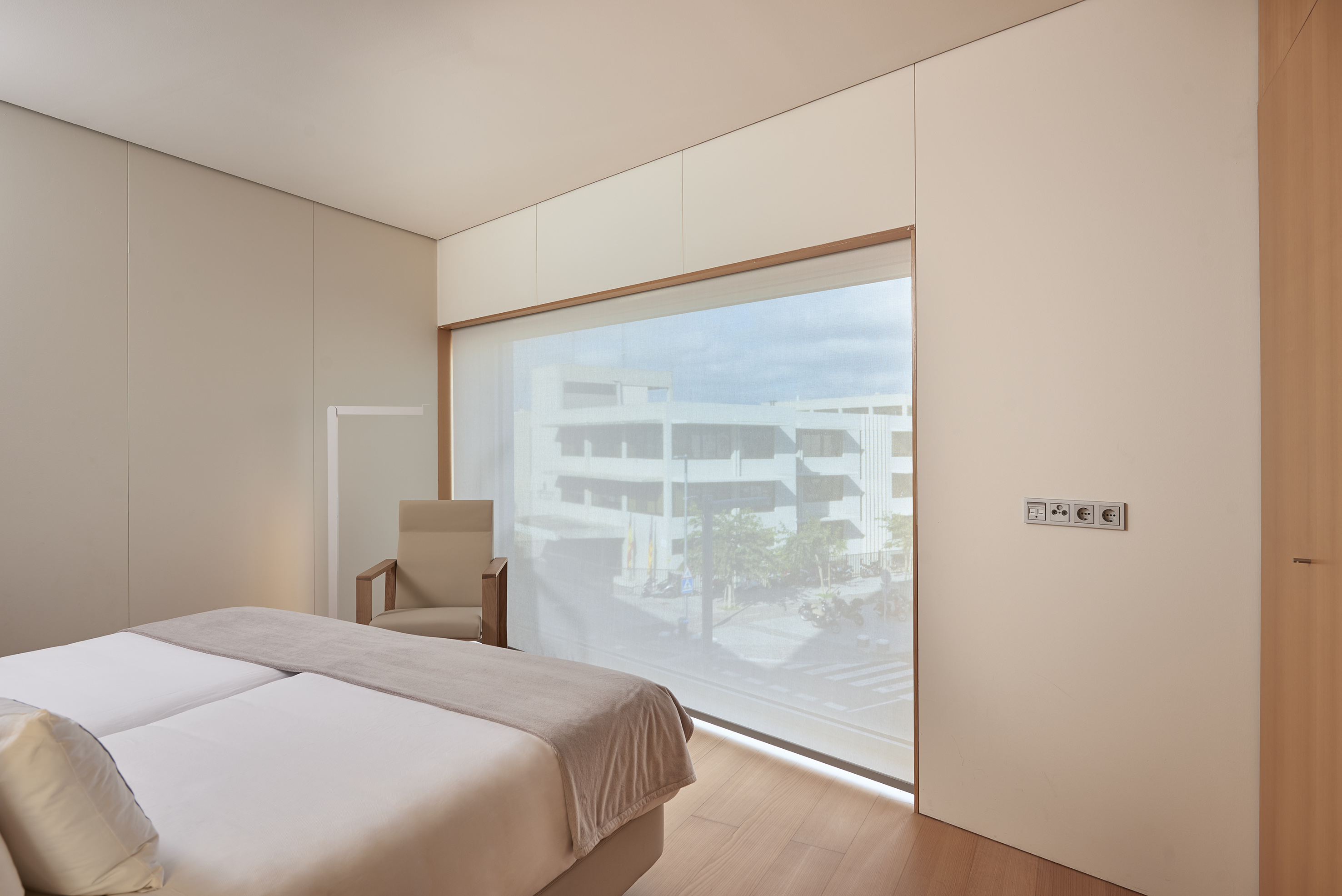 Minimalist twin bedroom with sheer blind window showing city buildings.