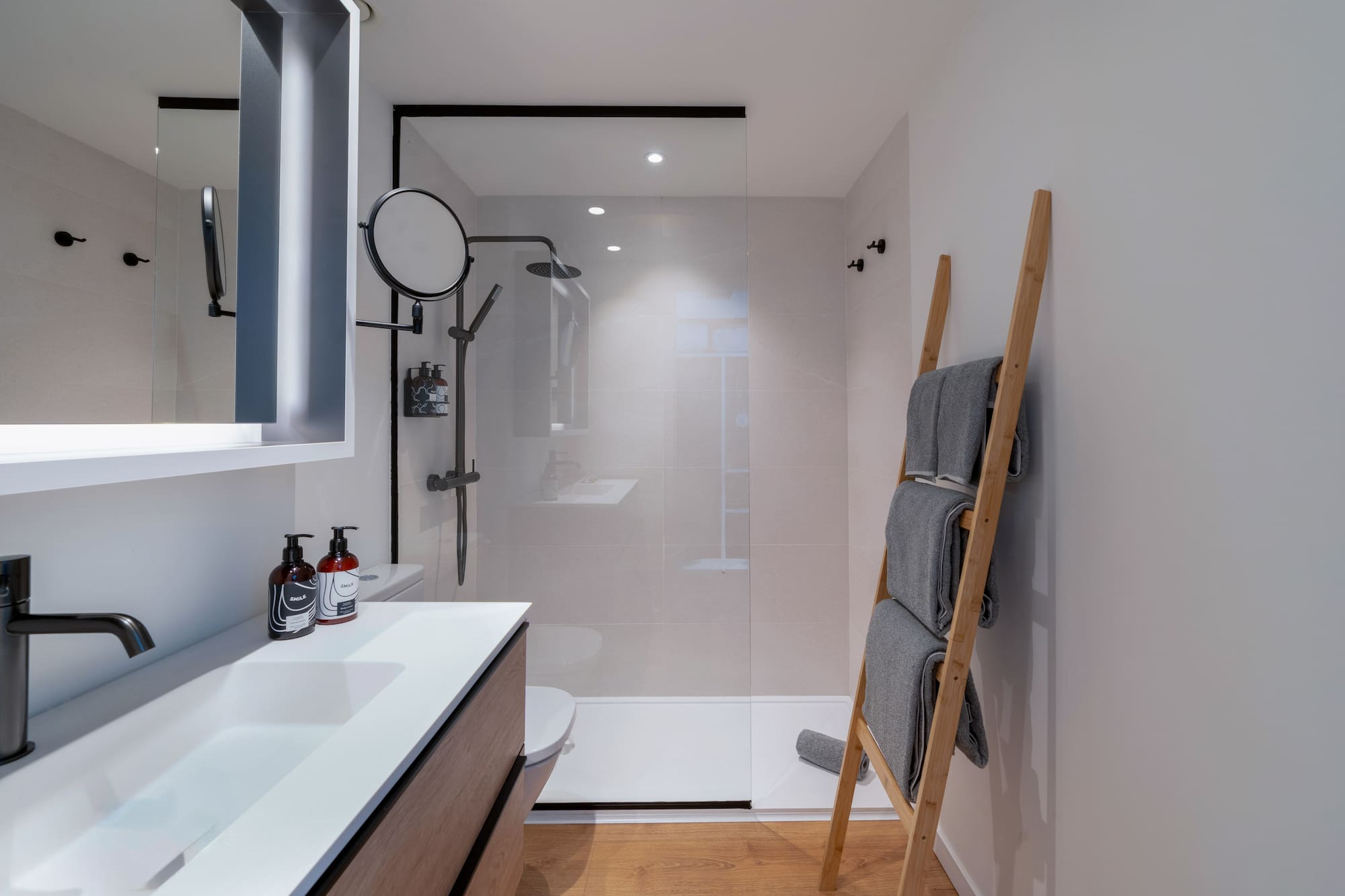 Baño moderno con ducha, lavabo, toallero de escalera y suelo de madera.