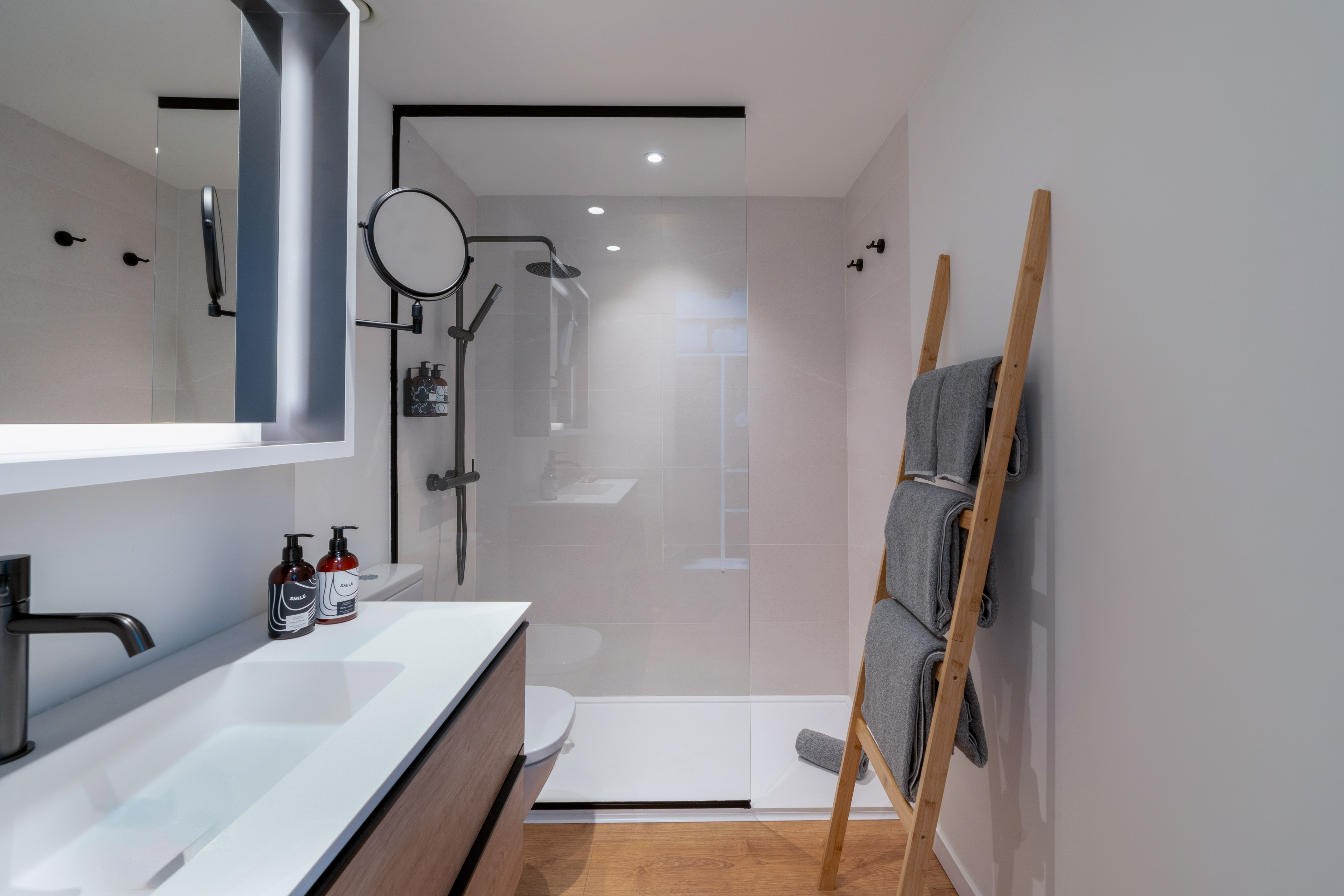 Baño moderno con ducha, lavabo, toallero de escalera y suelo de madera.