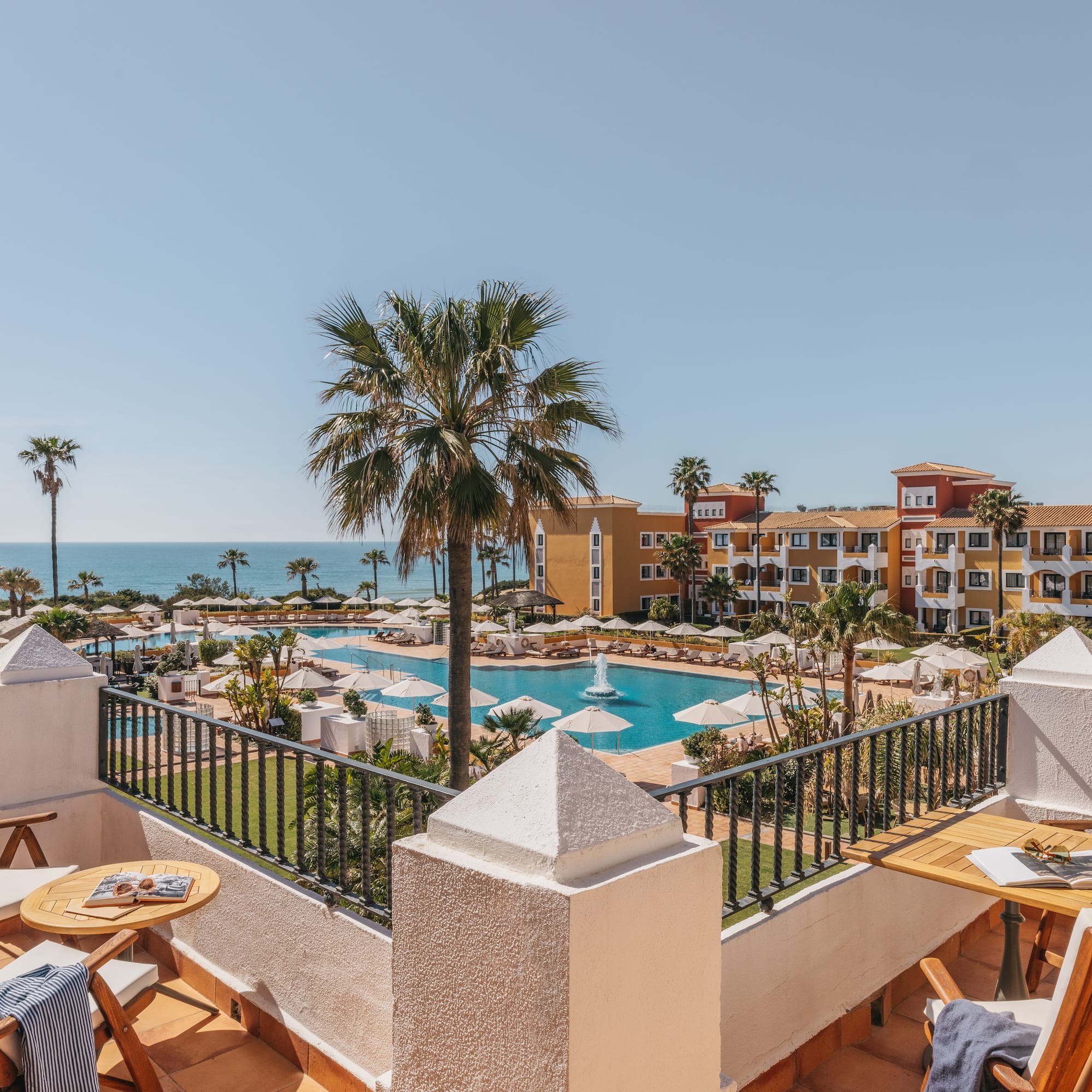 Balcony view of luxury resort with large swimming pool, palm trees, and ocean.