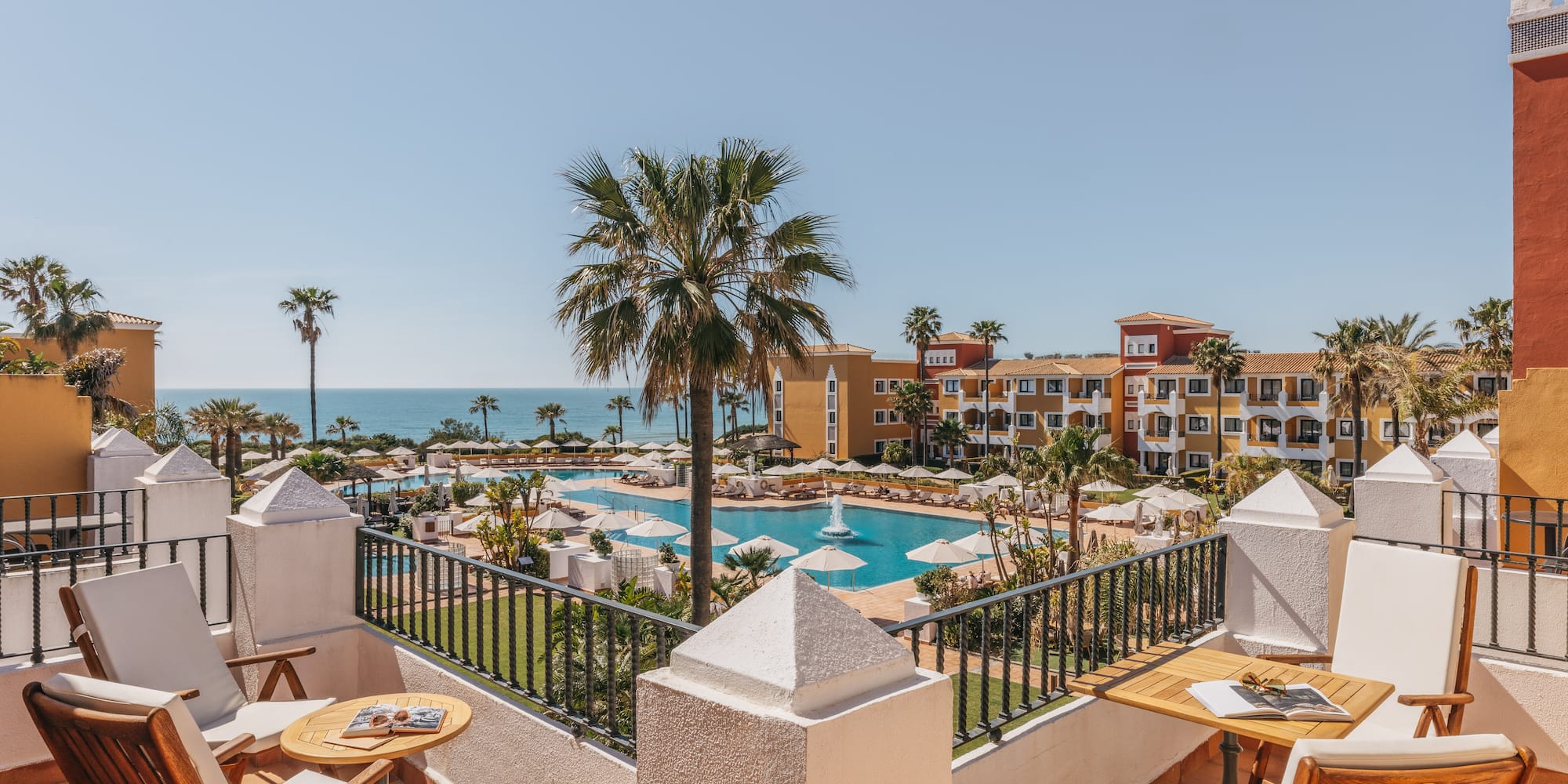 Balcony view of luxury resort with large swimming pool, palm trees, and ocean.