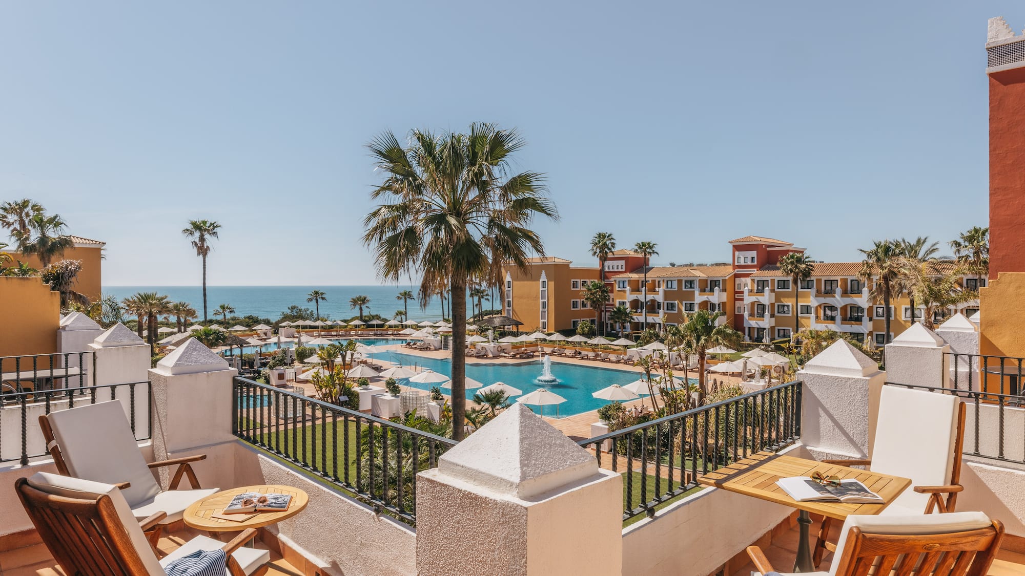 Balcony view of luxury resort with large swimming pool, palm trees, and ocean.