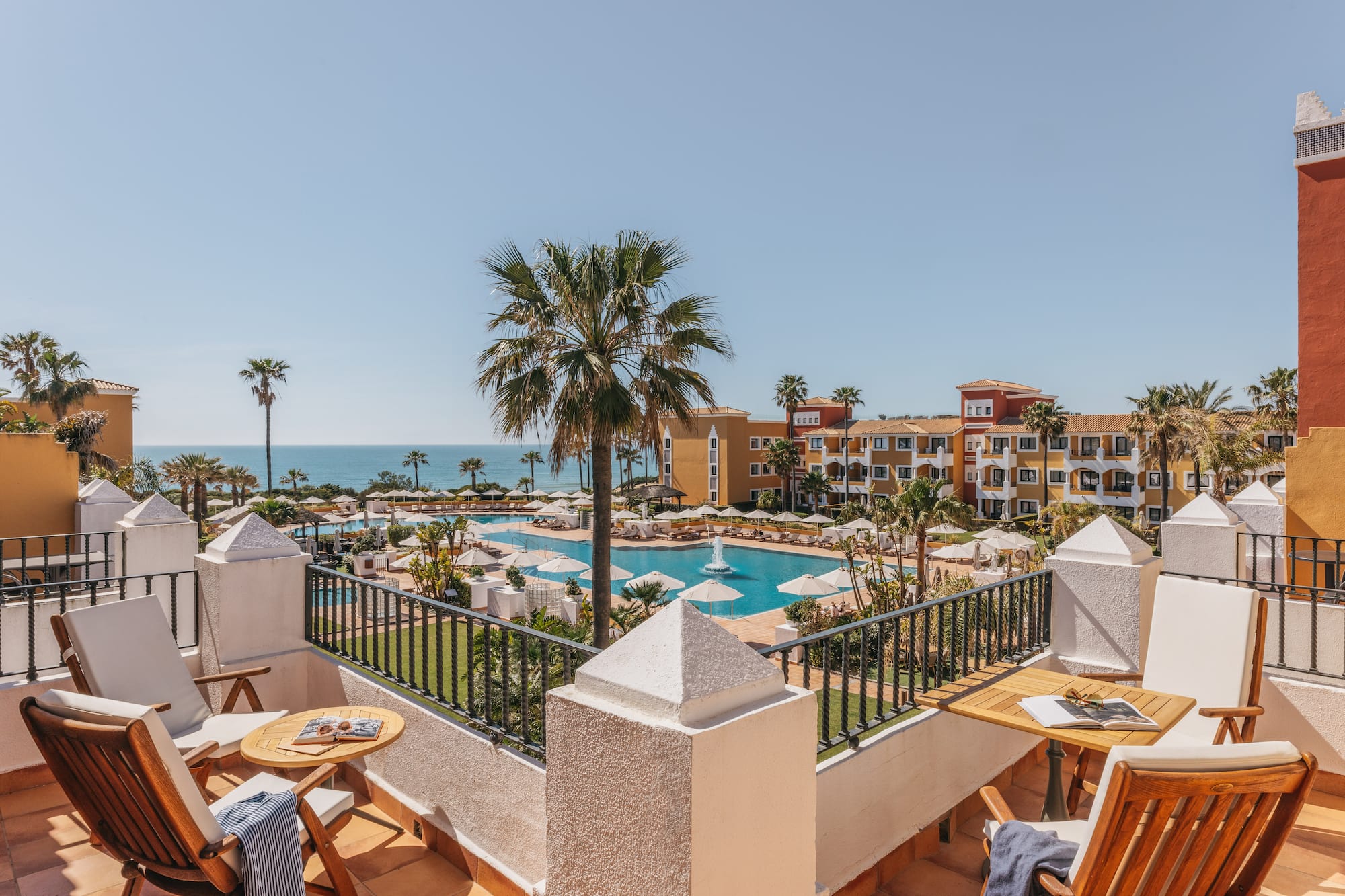 Balcony view of luxury resort with large swimming pool, palm trees, and ocean.