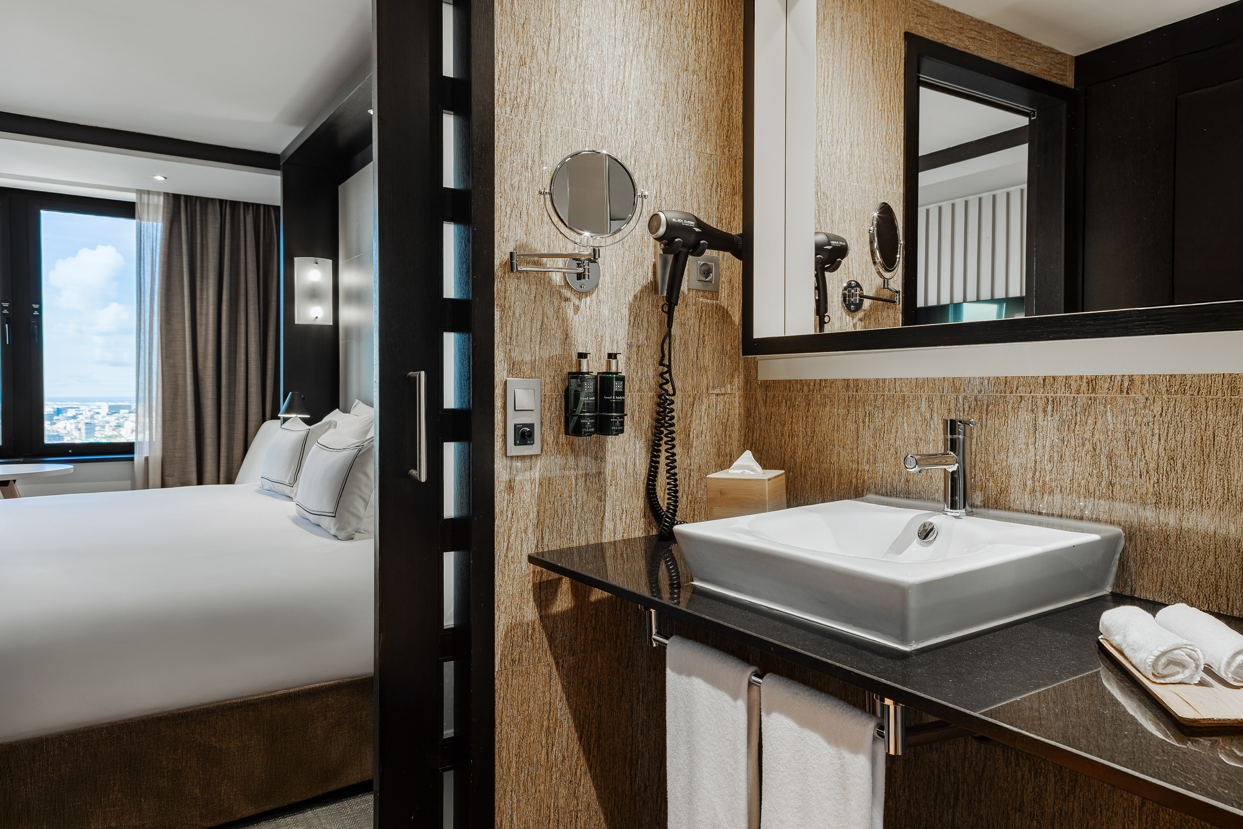 Modern hotel bathroom vanity with sink, mirror, hairdryer, and bedroom view.