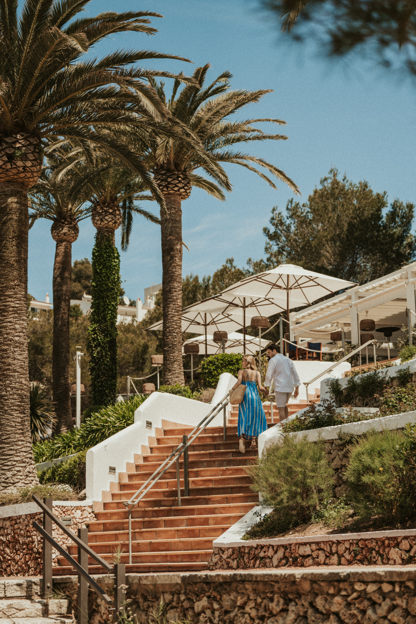 Tropical resort: couple ascending terracotta stairs past palm trees to outdoor dining.