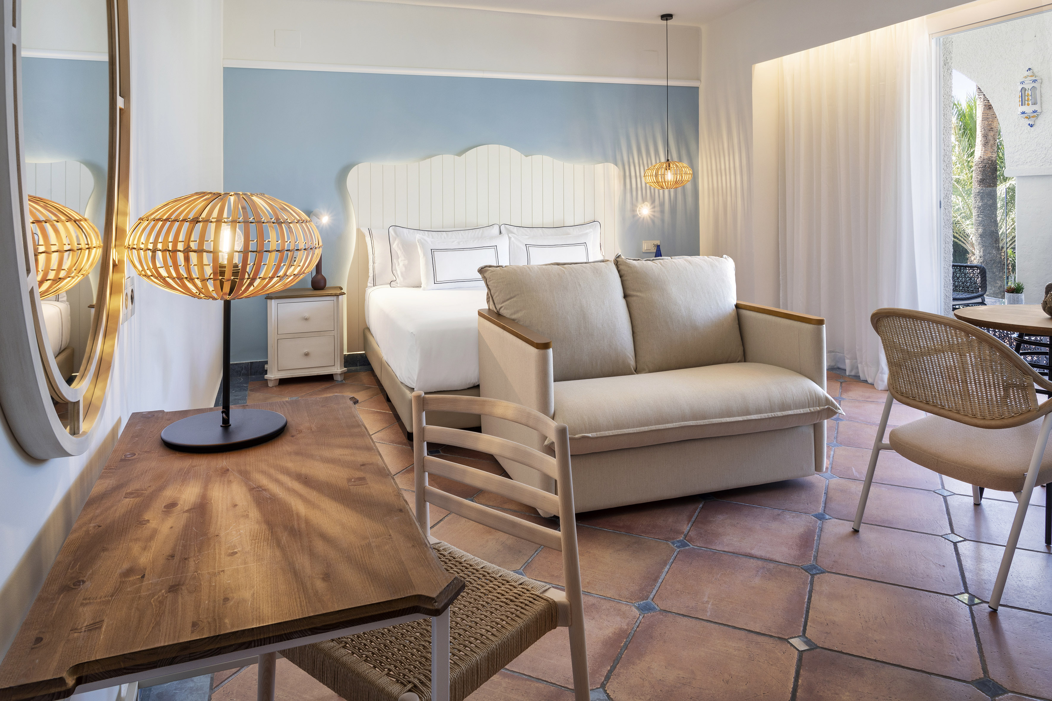 Bright hotel room with white bed, blue wall, sofa, and terracotta tiled floor.