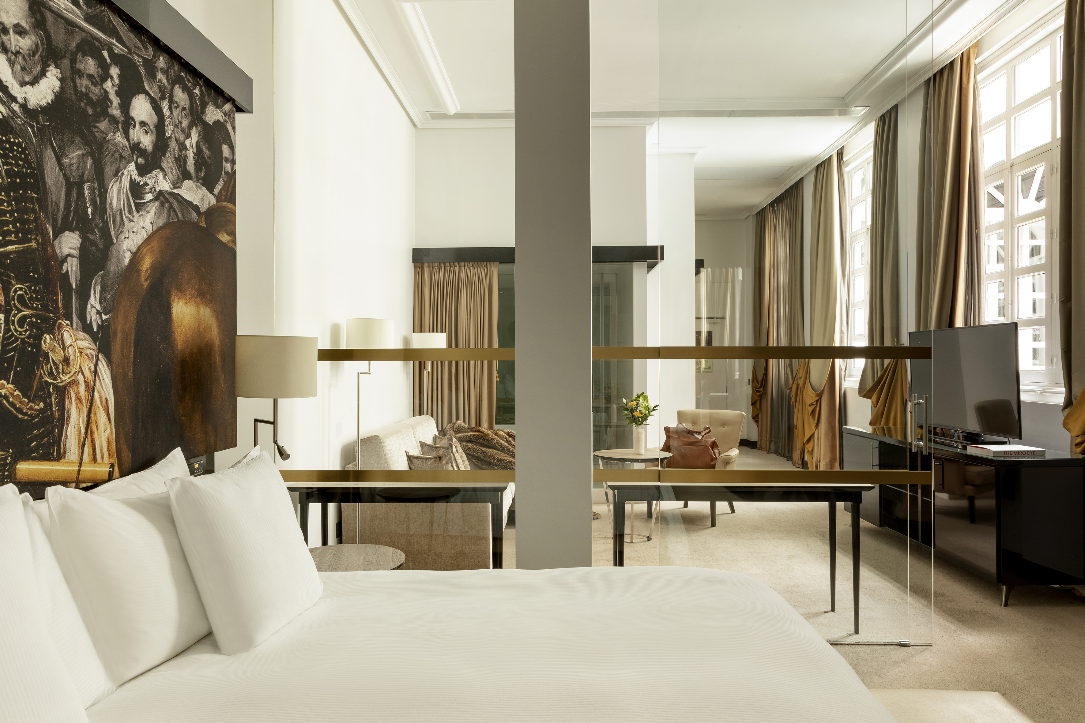 Elegant hotel suite with white bed, artwork mural, and living area behind glass.