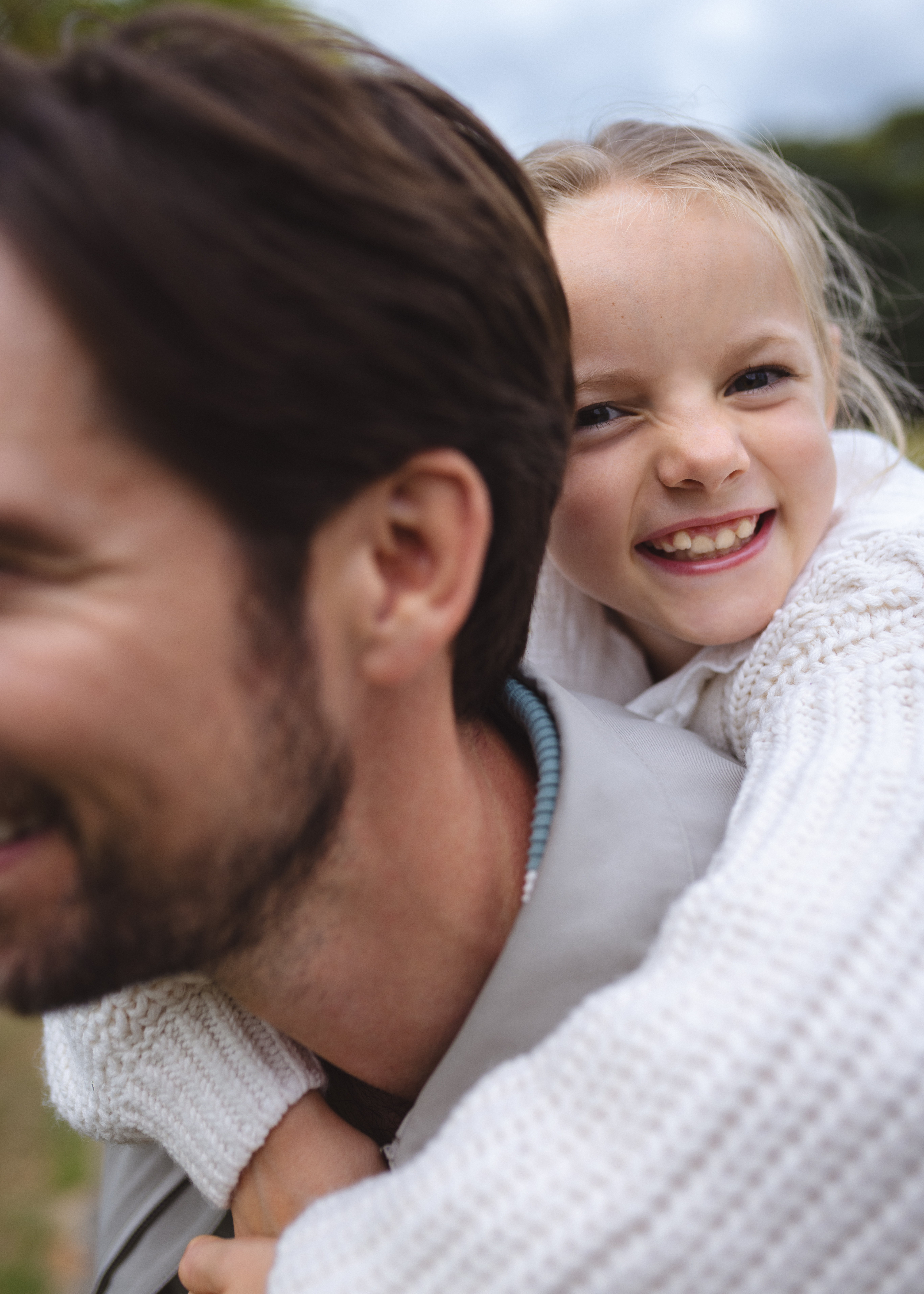 Happy blonde girl piggybacking father, both smiling brightly outdoors.