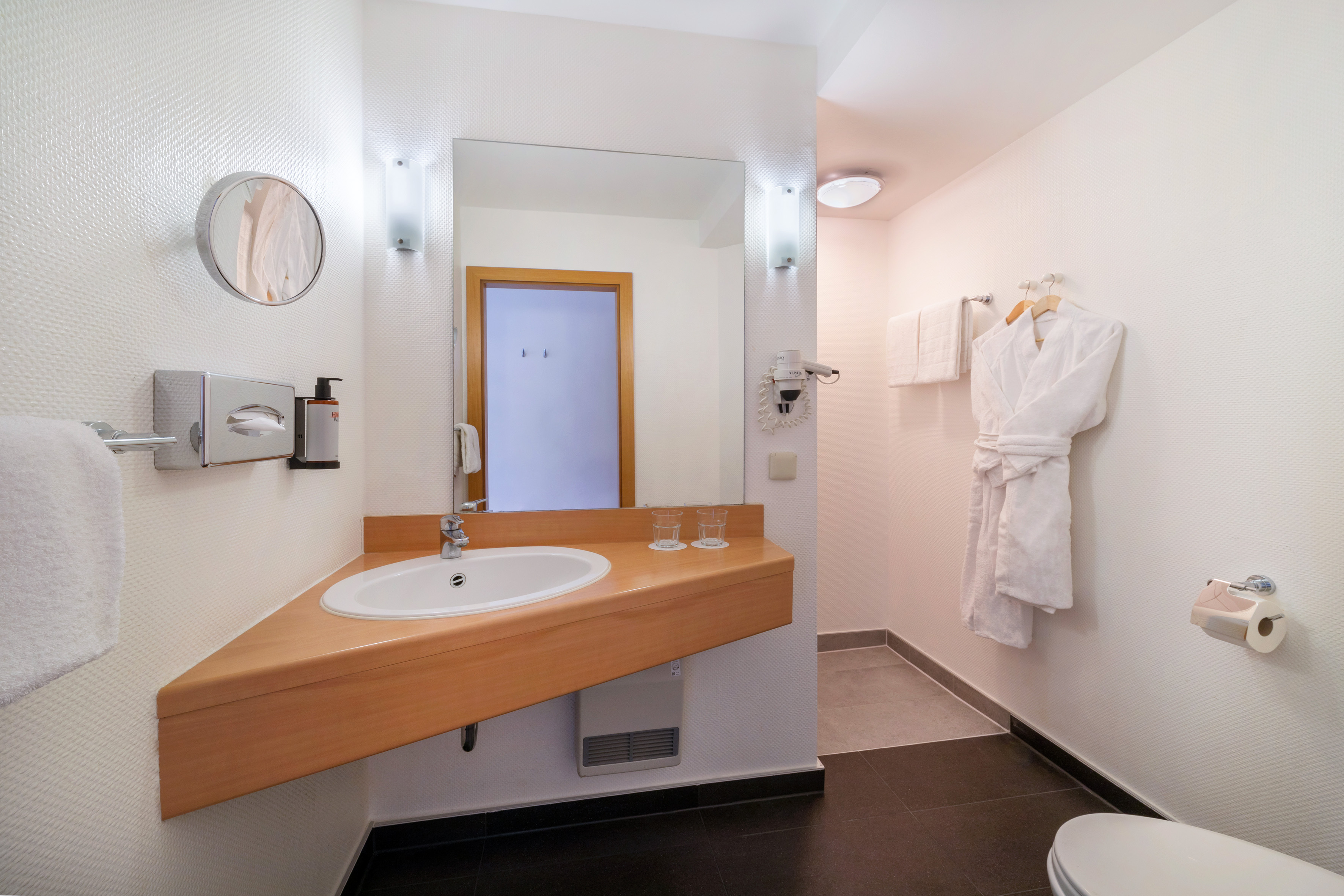 Modern, bright hotel bathroom with white sink, large mirror, wooden vanity, and bathrobe.