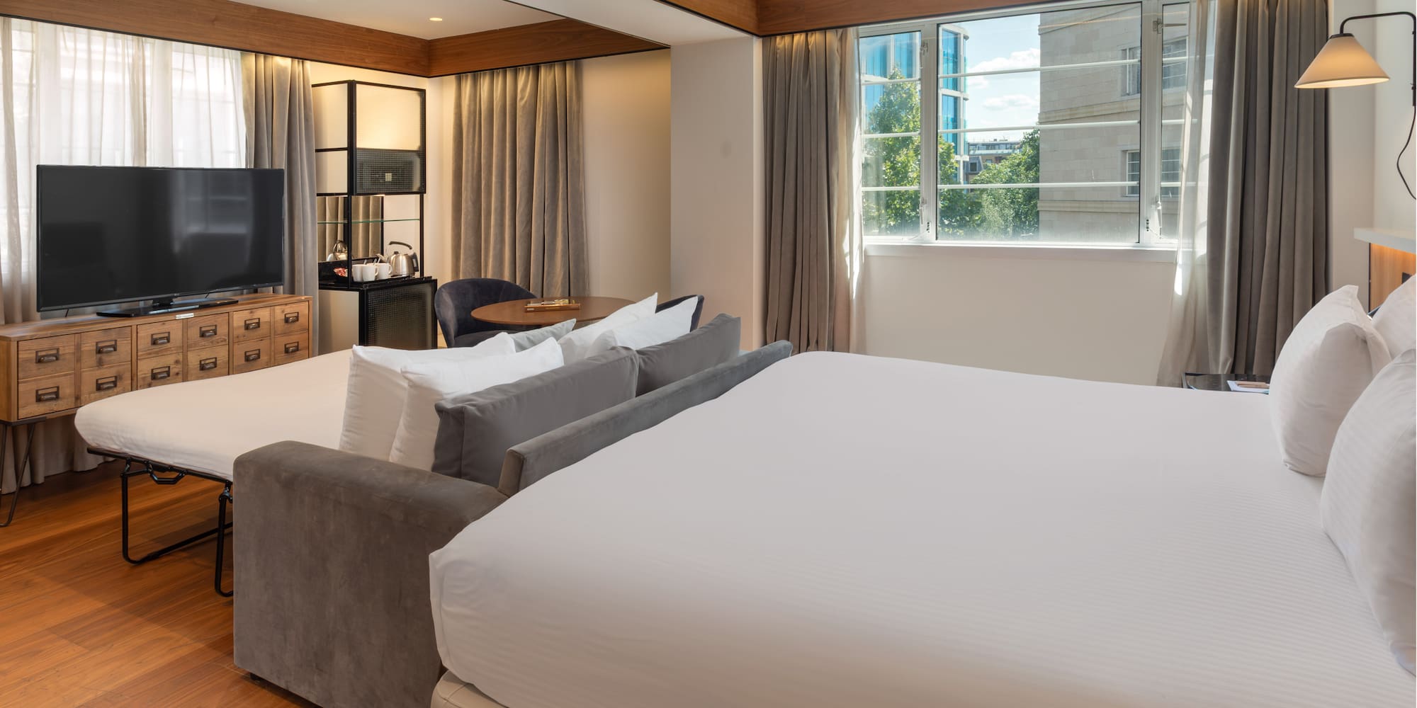 Modern hotel room with king bed, sofa bed, TV, wood floor, and city view window.