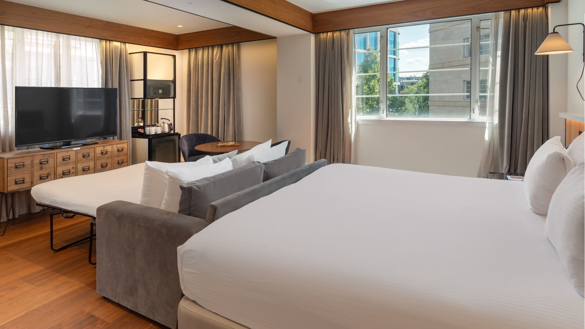Modern hotel room with king bed, sofa bed, TV, wood floor, and city view window.