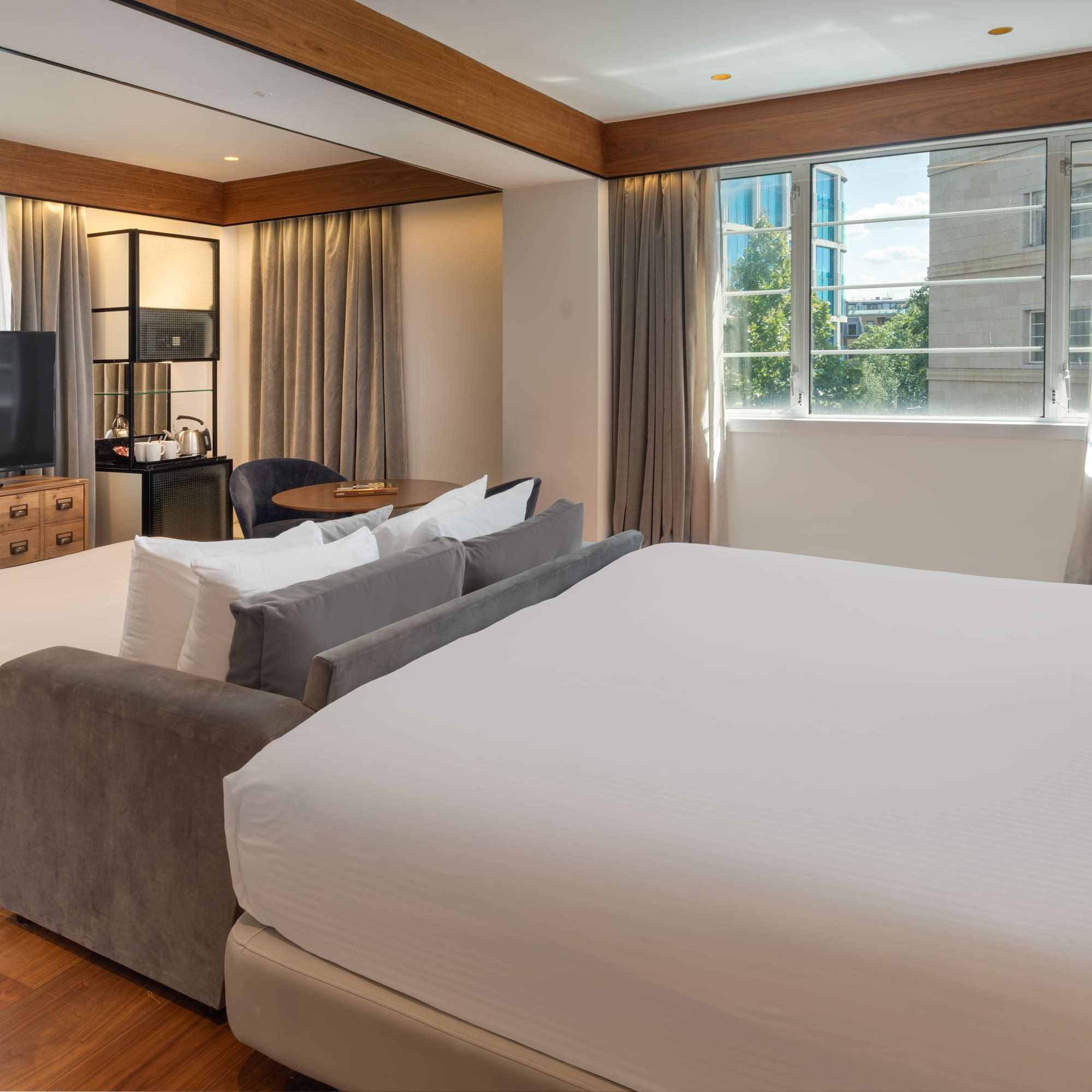 Modern hotel room with king bed, sofa bed, TV, wood floor, and city view window.