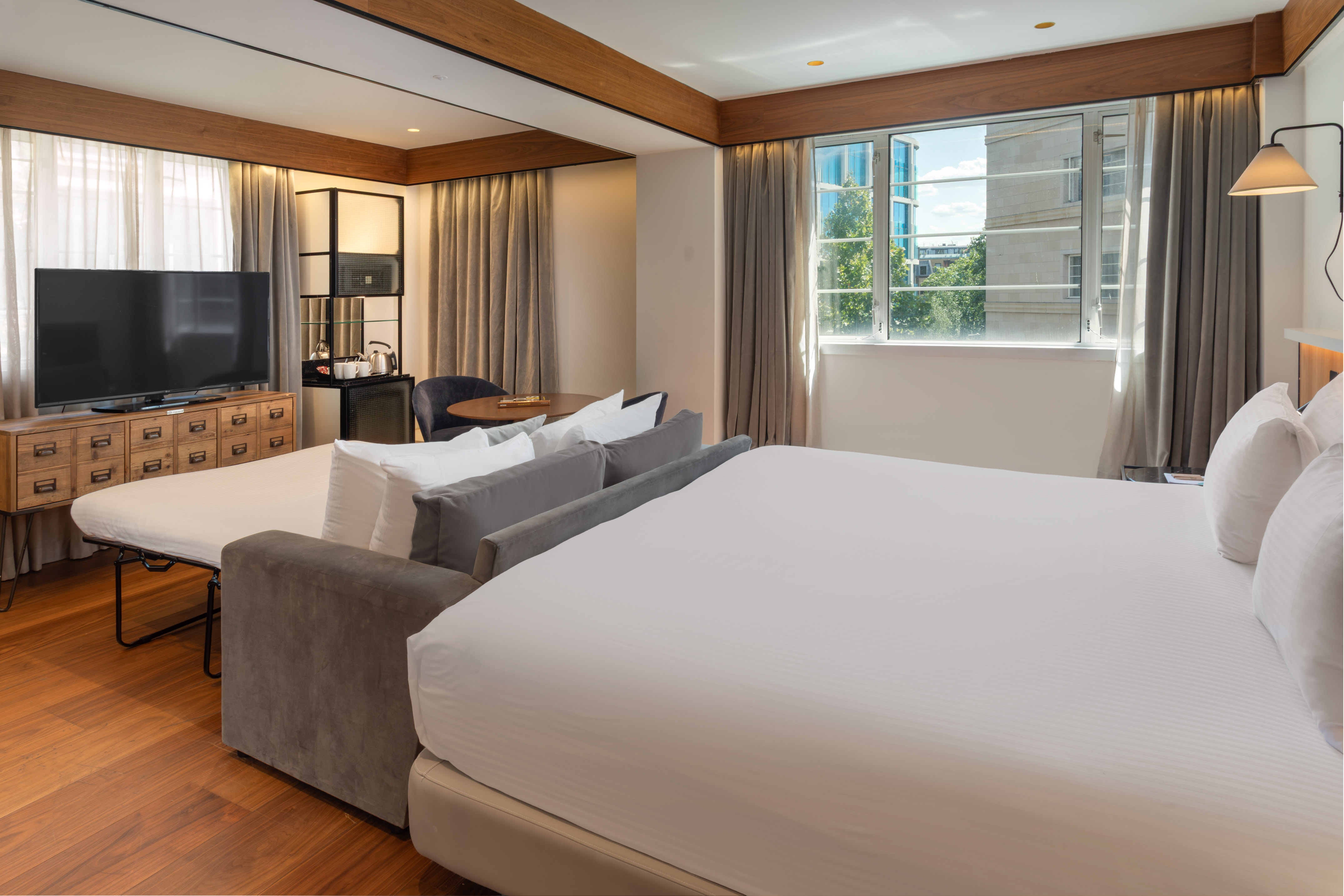 Modern hotel room with king bed, sofa bed, TV, wood floor, and city view window.
